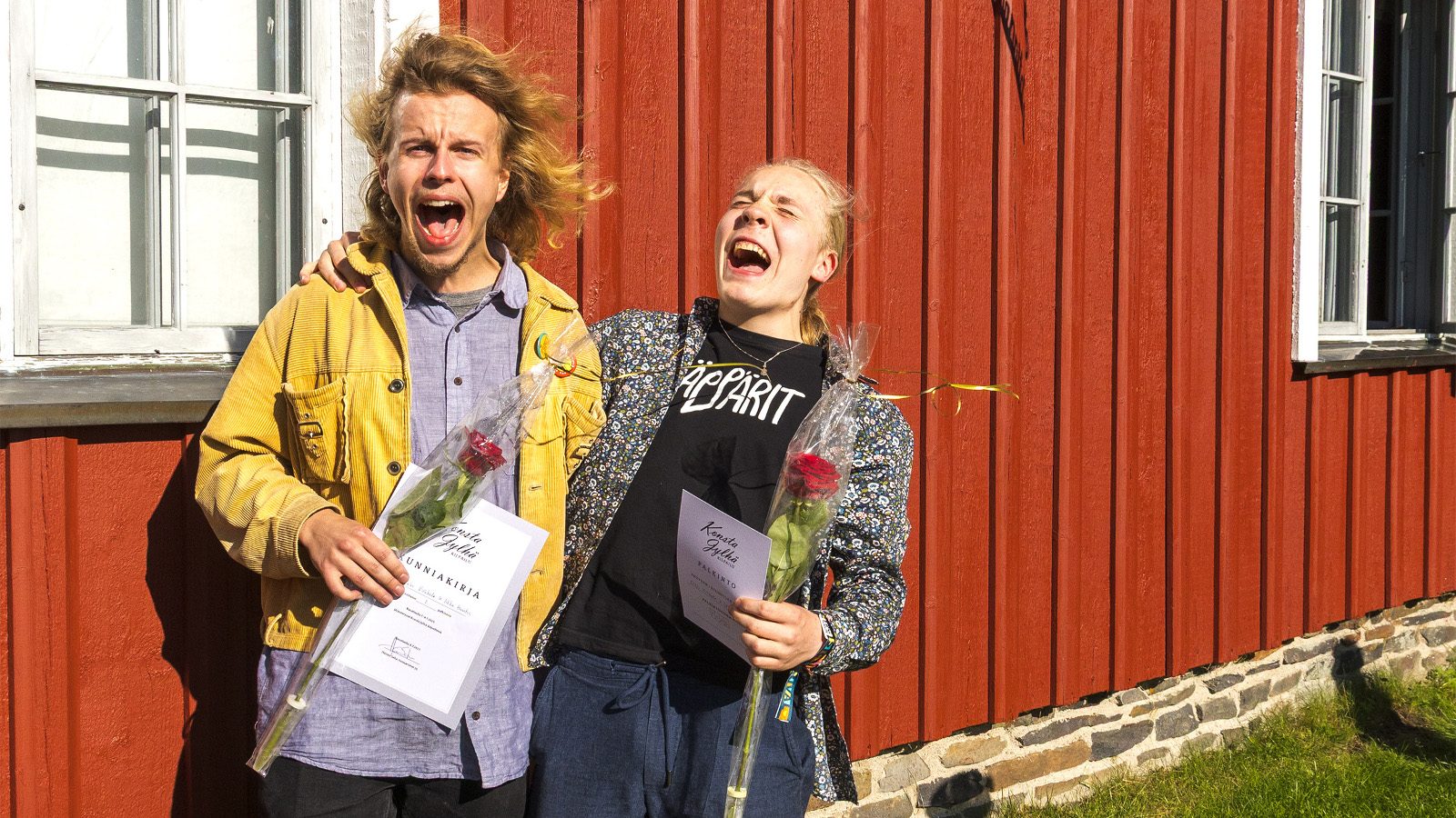 Kuvassa ovat Erkki Virkkala keltaisessa takissa. Oikealla hänen vieressään on Iikka Huntus harmaassa takissa ja mustassa t-paidassa. Molemmat ovat innoissaan. Takana on punaista talon seinää ja valoinen ikkuna. 