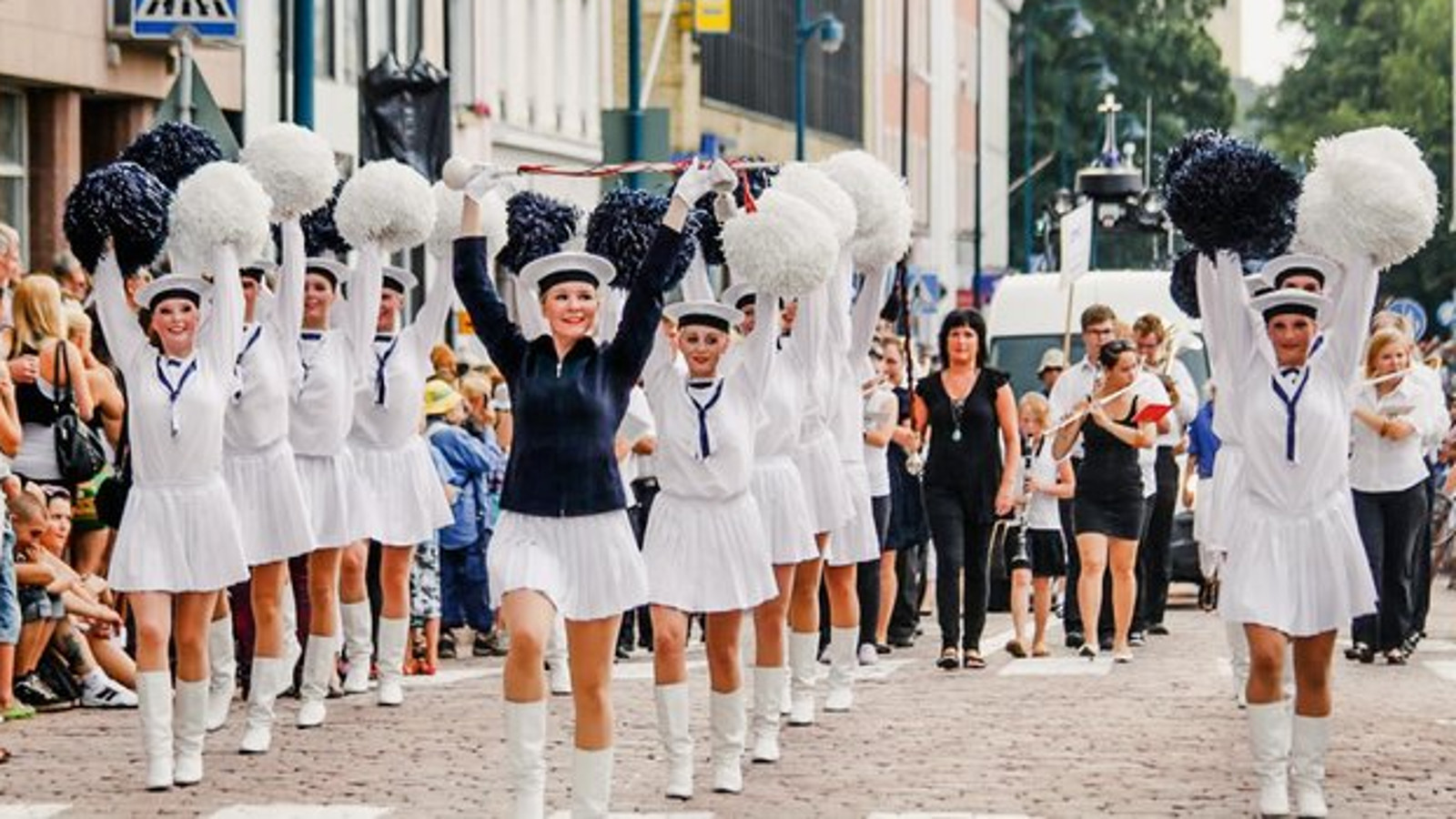 Kuvassa on kolmessa rivissä nuoria naisia marssimassa merimies-henkisissä, valkoisissa puolihamessa  ja puseroissa.  Heillä on kädet ylhääällä ja käsissä siniset tai valkoiset paperiset viuhkat.  Kadun molemmin puolin on yleisöä kadunvarsissa.