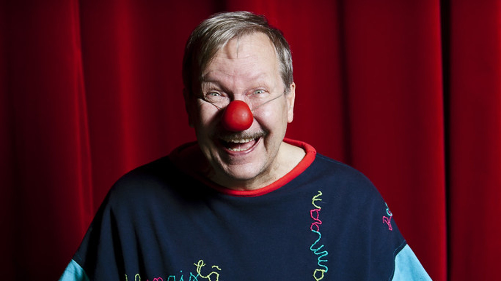 Kuvassa on Erkki Saarela puolivartalokuvassa. Hänellä on tumma college-paita ja pyöreä, punainen tekonenä.  Taustalla on tummanpunainen verho.