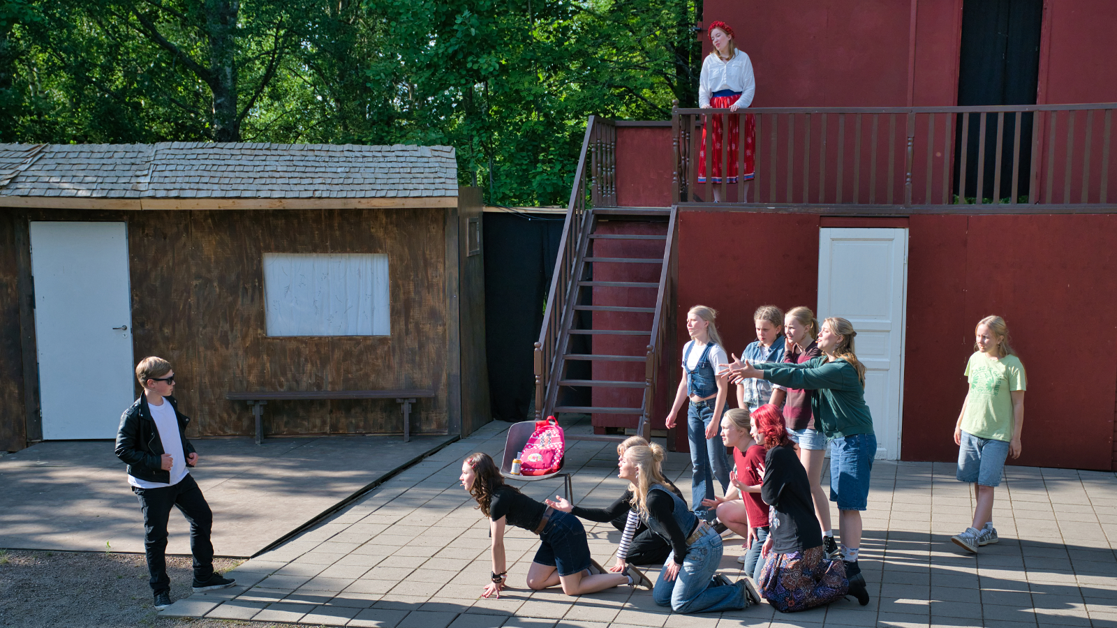 Kuvassa on kohtaus, jossa vasemmalla seisova mies keskustelee näyttämöllä olevan joukon kanssa ja lavasteen parvekkeelta katselee yksi nainen. Vasemmalla on ruskea tupa ja oikealla korkea lavaste.
