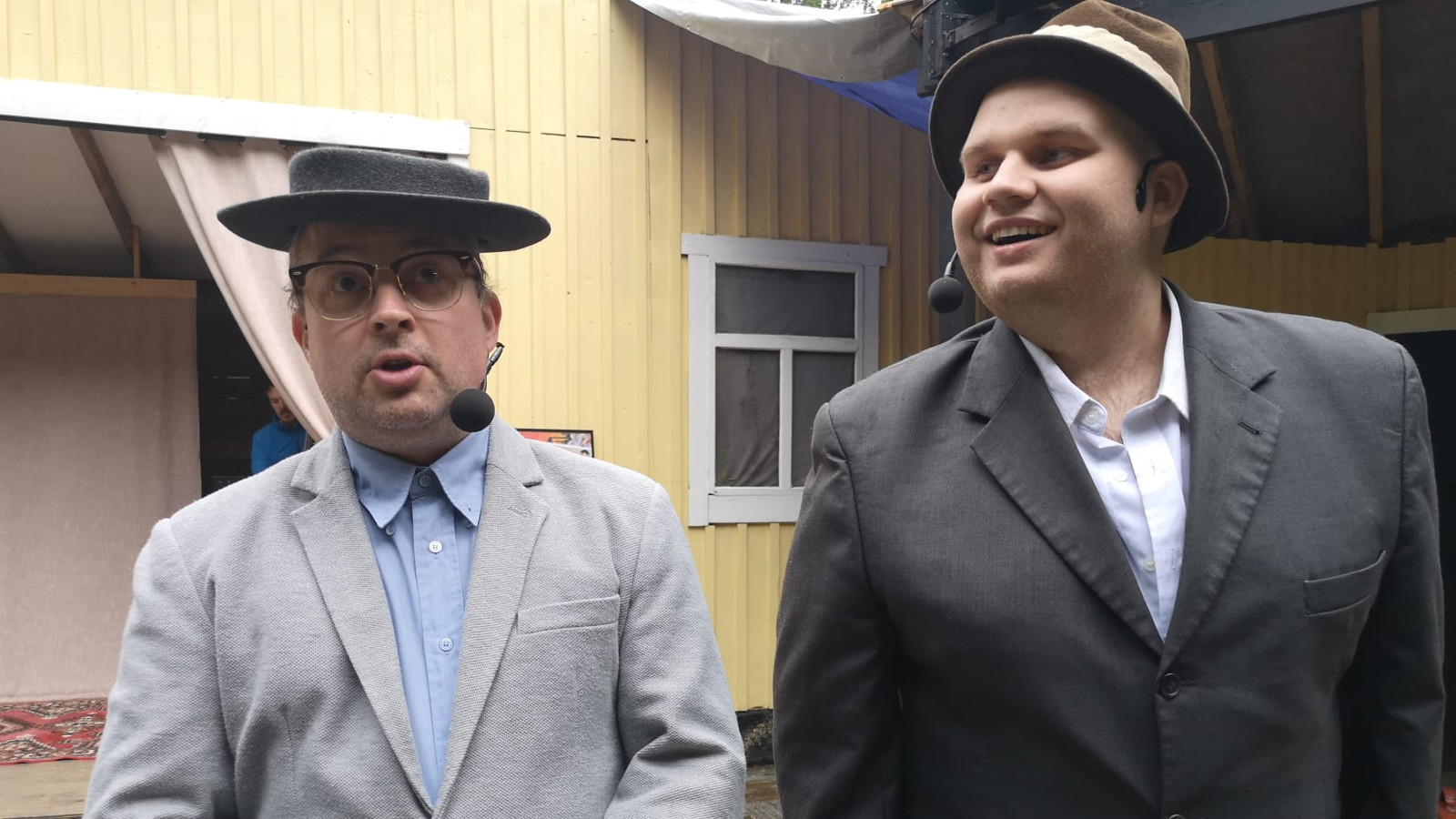 Kuvassa on kaksi miestä puolivartalokuvassa vasemmalla mies vaaleassa takissa ja oikealla pidempi mies tummassa takissa. Taustalla on keltaista talon seinää.