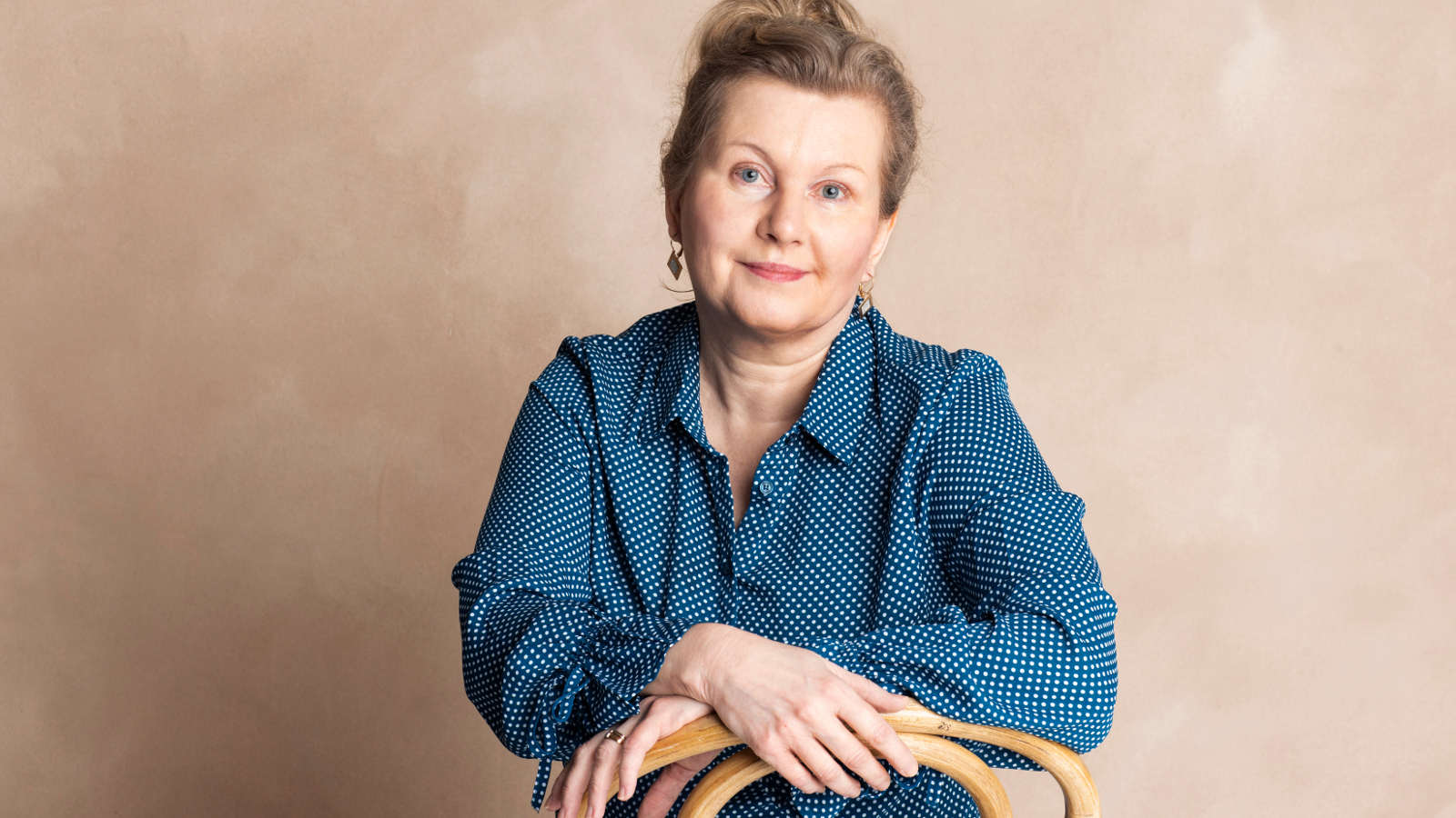 Kuvassa Tuula Salminen istumassa ja hänellä on käsivarret ristikkäin edessään. Hänellä on sininen pusero päällään. Tausta on ruskehtava.
