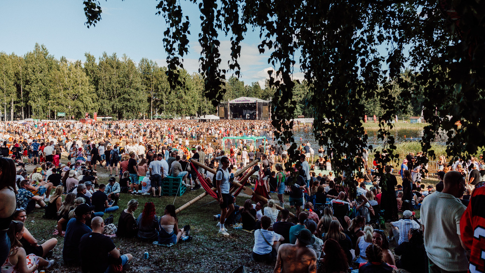 Kuvassa on kesäisessä säässä tuhansia ihmisiä tasaisella maalla ja taustalla on metsää. Edessä näkyy koivun oksia.