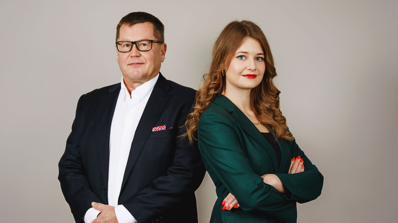 Kuvassa on vasemmalla Jukka Savolainen mustassa takissa ja valkoisessa paidassa. Oikealla on vinottain seisoo Iida Hallikainen ja hänellä on tumman vihreä jakku.  Kuvassa ovat he ovat puolivartalokuvassa. 