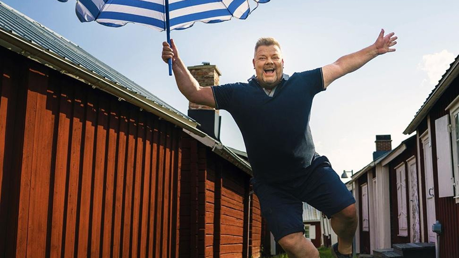 Kuvassa on Jari Sillanpää, joka hyppää sini-valkoinen päivänvarjo oikeassa kädessään. Hänellä on tumman sininen t-paita ja shortsit  päällään. Taustalla on katu, jossa on punaisia, matalia rakennuksia.