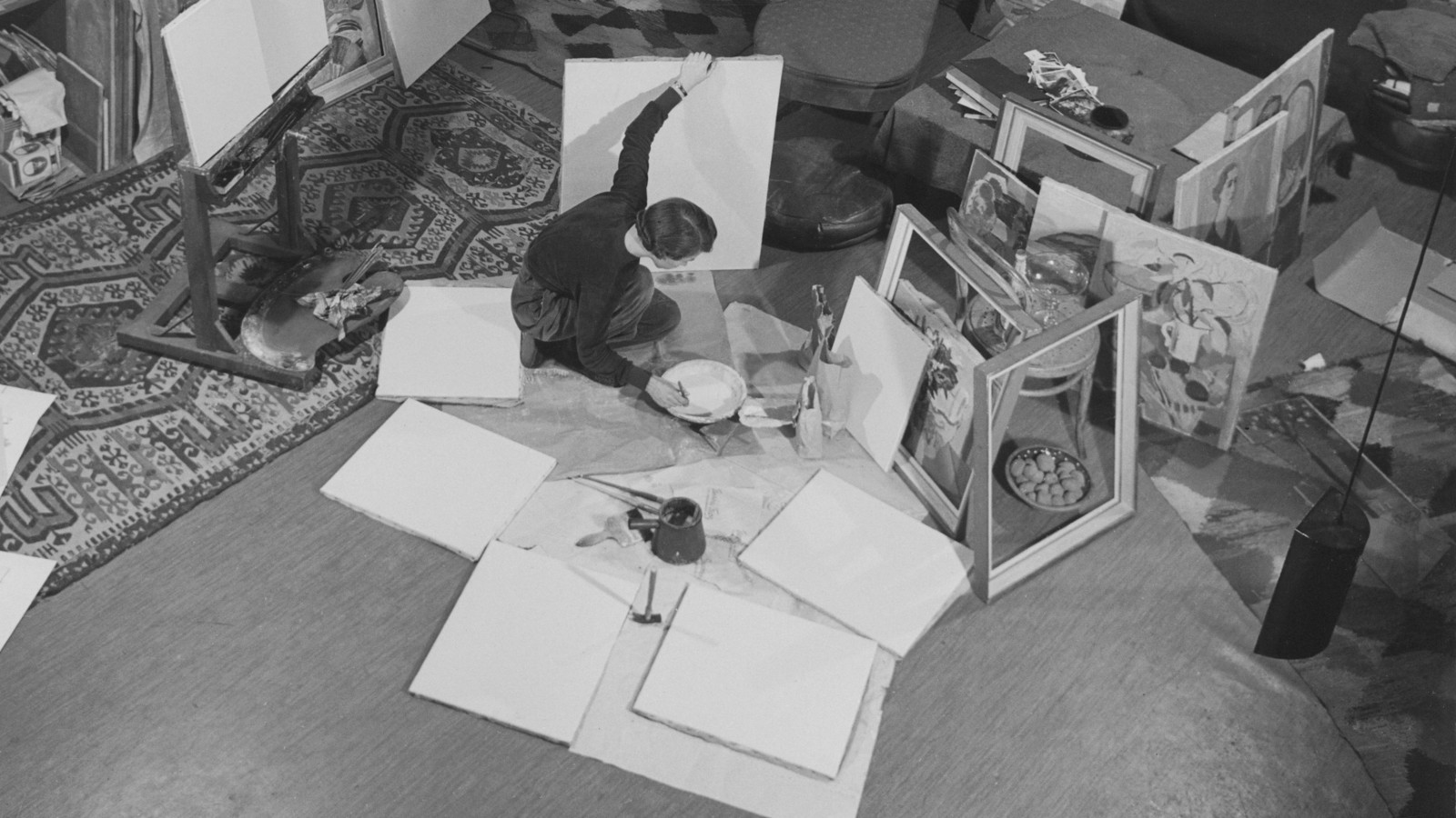Kuvassa on Tove Jansson kumartuneena lattialla maalaamaan. Lattialla on paljon piirrospaperia. Kuva on musta-valkoinen.