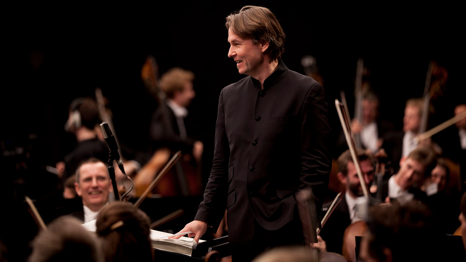 Kuvassa on Esä-Pekka Salonen ylempänä orkesterin keskellä ja hänellä on musta paita päällään. Tausta on musta.