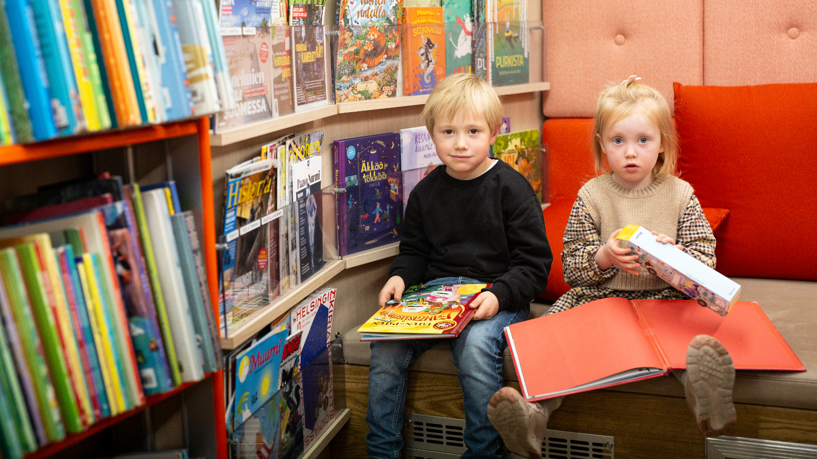 Kuvassa vasemmalla kirjahyllyä, jossa on värikkäitä lastenkirjoja. Vieressä oikealla sohvalla istuvat vasemmalla pikkupoika mustassa puserossa ja kirja sylissään. Hänen vieressään istuu pikkutyttö iso kirja avattuna sylissään.