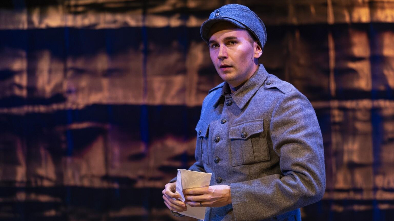 Kuvassa on harmaan sinisessä puvussa seisova sotilas, joka pitää kirjettä kädessään. Taustalla on ruskea seinä.