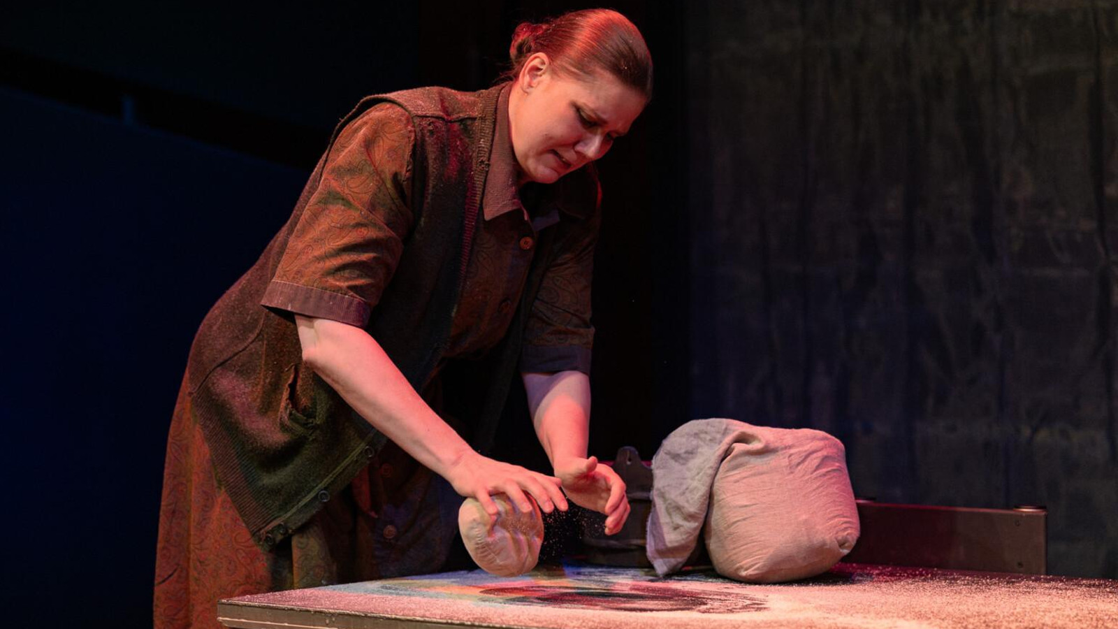 Kuvassa on nainen leopomassa ruisleipää hieman sivuttain. Hnellä on ruskea mekko päällään. Pöydällä vieressä pieni jauhosäkki.