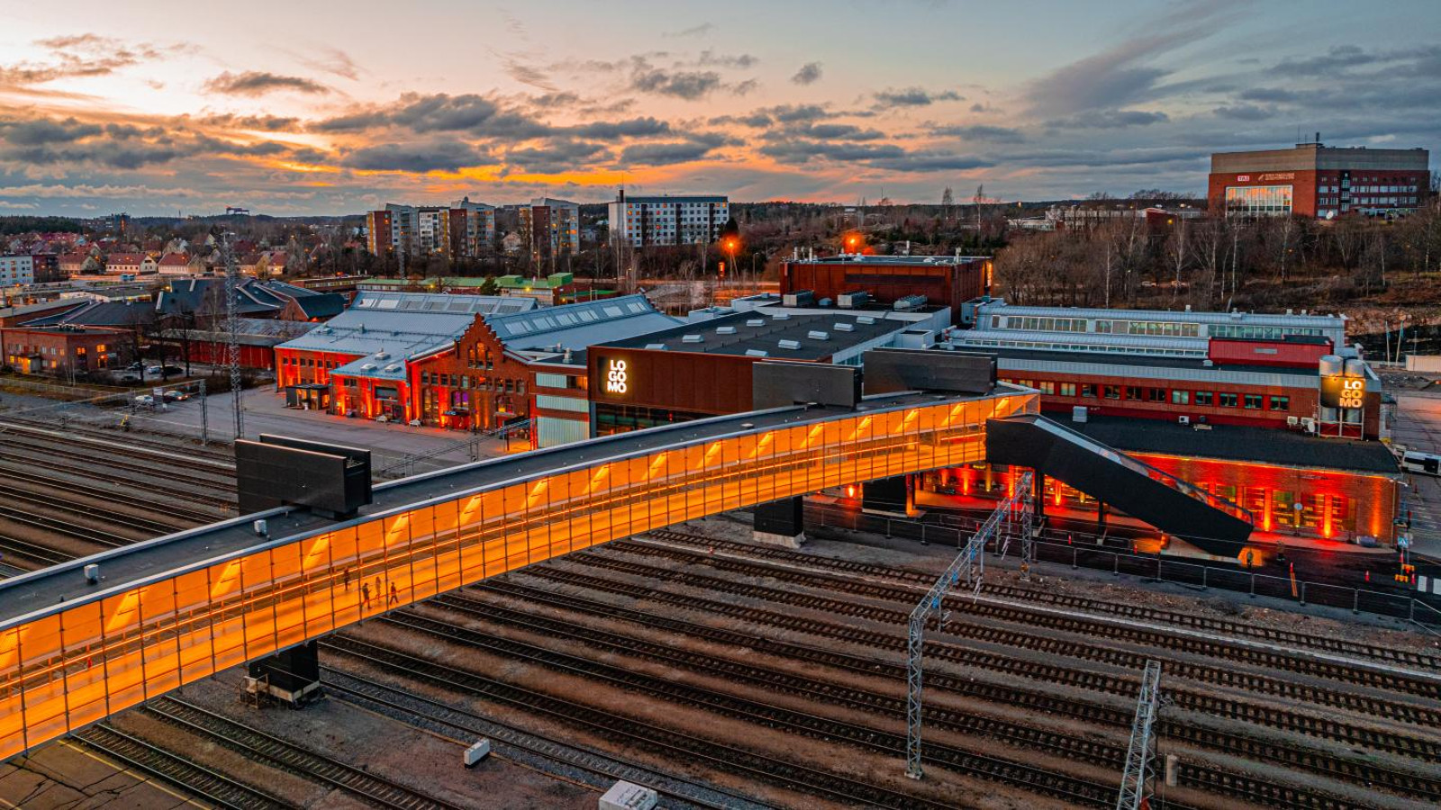 Kuvassa on Turun Logomon rakennukset ja sen ympäristöä iltahämärässä. Alaosassa on valaistu silta, joka on rautieraiteiden yläpuolella.