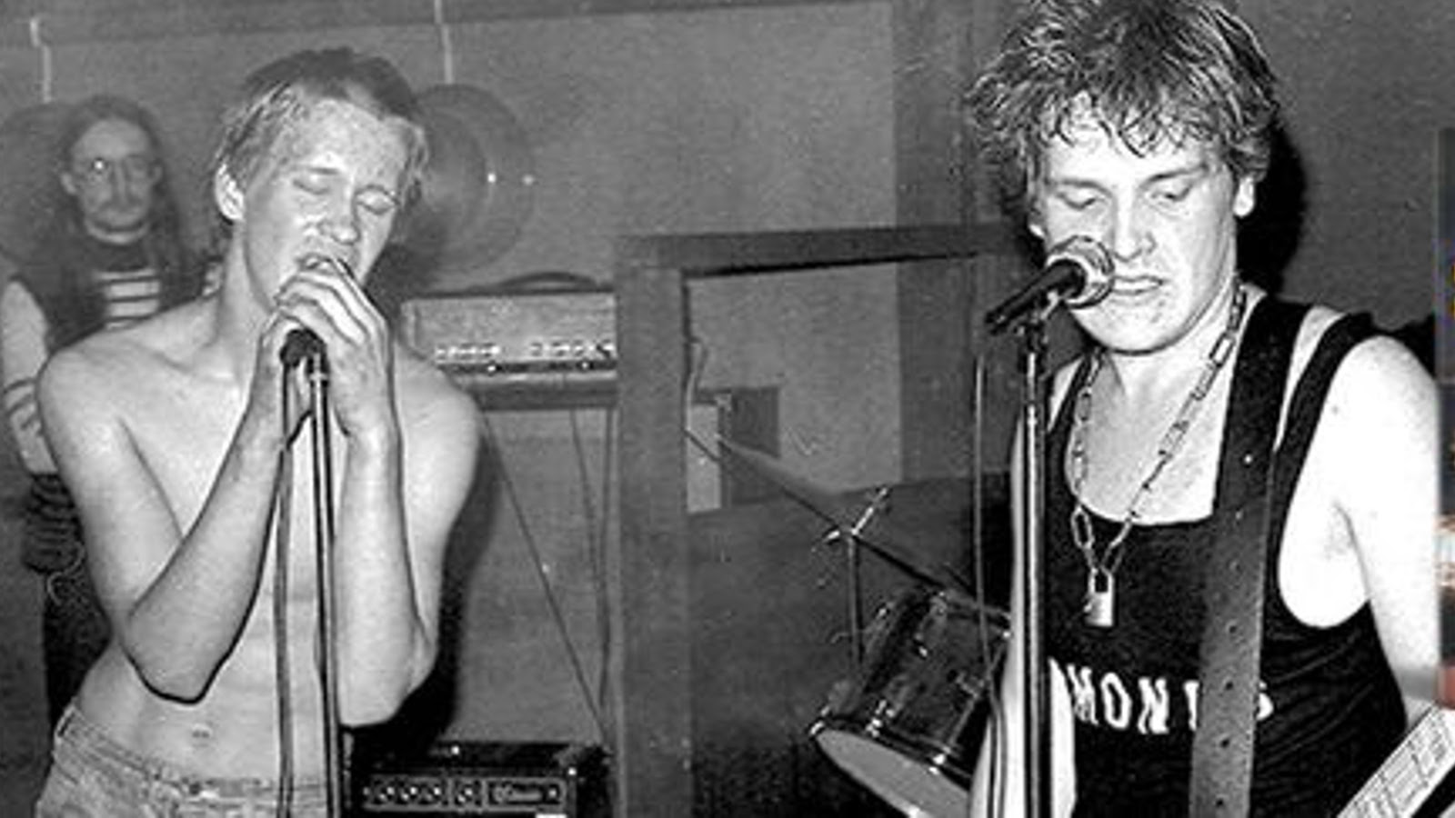 Kuvassa on vasemmalla Aku Syrjä laulamassa ilman paitaa ja oikealla on Mikko Saarela mustassa hihattomassa t-paidassa soittamassa kitaraa ja laulamassa mikrofoni edessään. Kuva on musta-valkoinen.