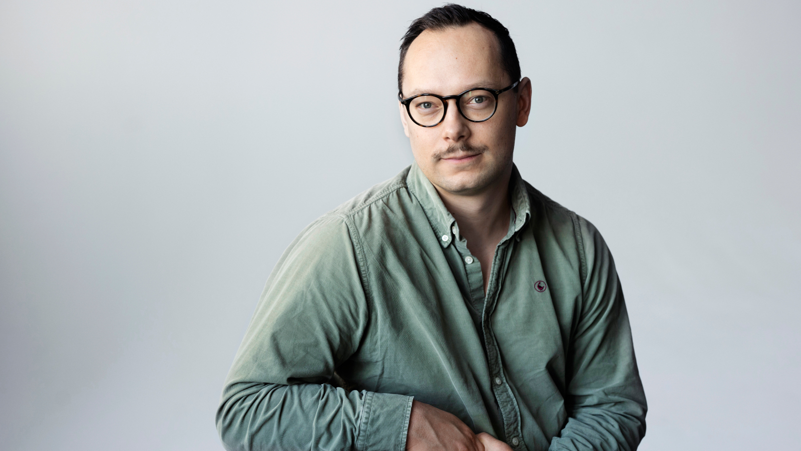 Kuvassa on Paavo Teittinen istumassa ja nojaa oikeaan kätensä. Hänellä on vihertävä paita. Tausta on harmaa.