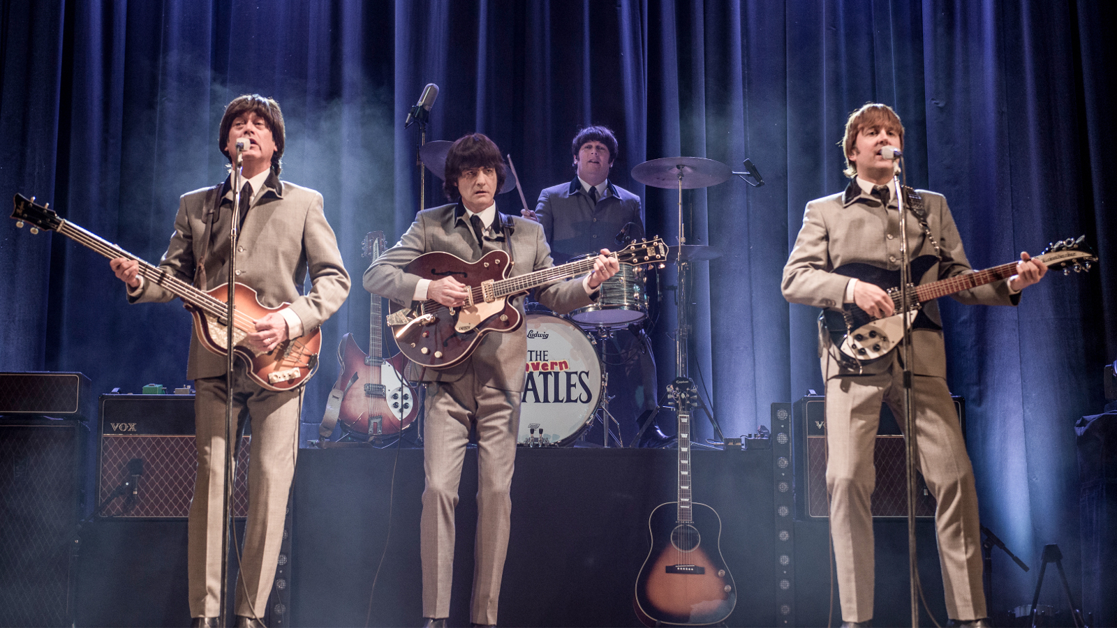 Kuvassa ovat The Beatles-tribuuttibändin kolme jäsentä edessä kitaroineen ja heillä on vaalean ruskeat puvut päällään. Taustalla on rumpali. Takana on tumman sininen verho.