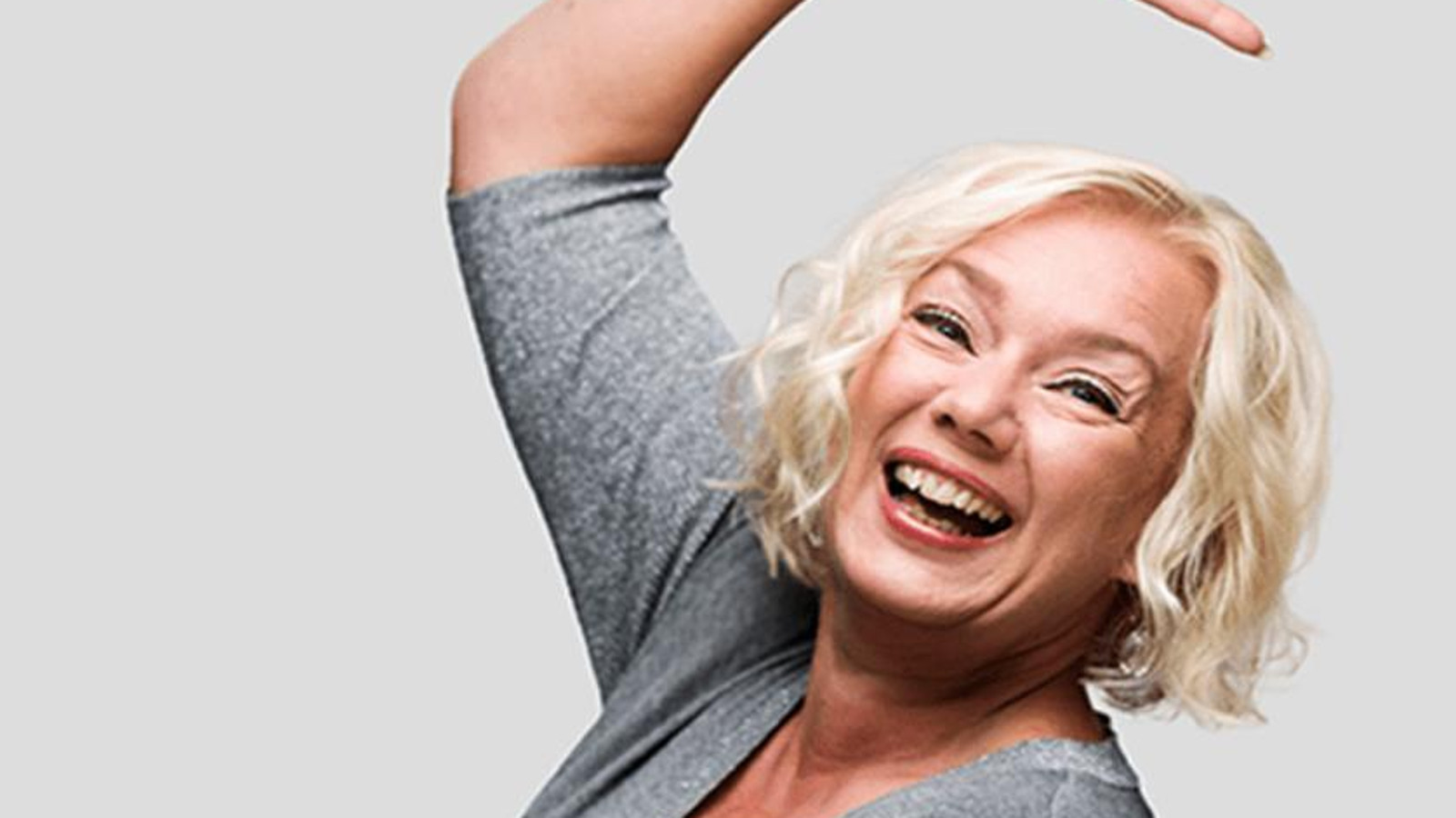 Kuvassa on Sari Siikander iloisesti oikea käsi ylös kohotettuna. Hänellä on harmaa pusero päällään ja vaaleat hiukset.