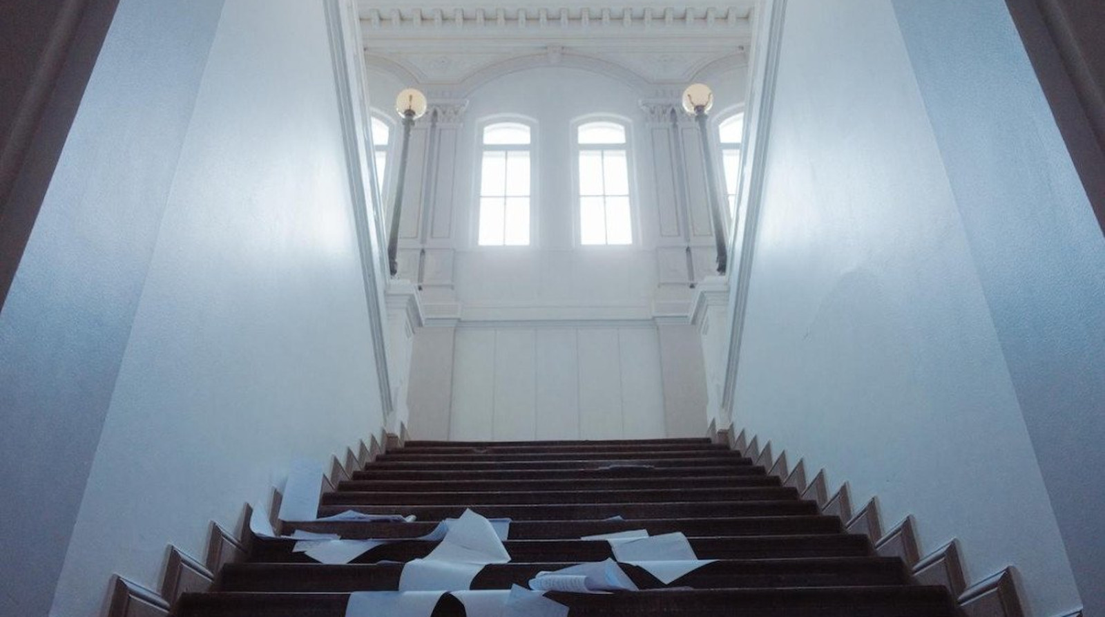Kuvassa on valkoinen ylöspäin menevä rappukäytävä, jossa on mustilla portailla valkoisia papereita. Ylhäällä on kaksi korkeaa ruutuikkunaa korkealla.