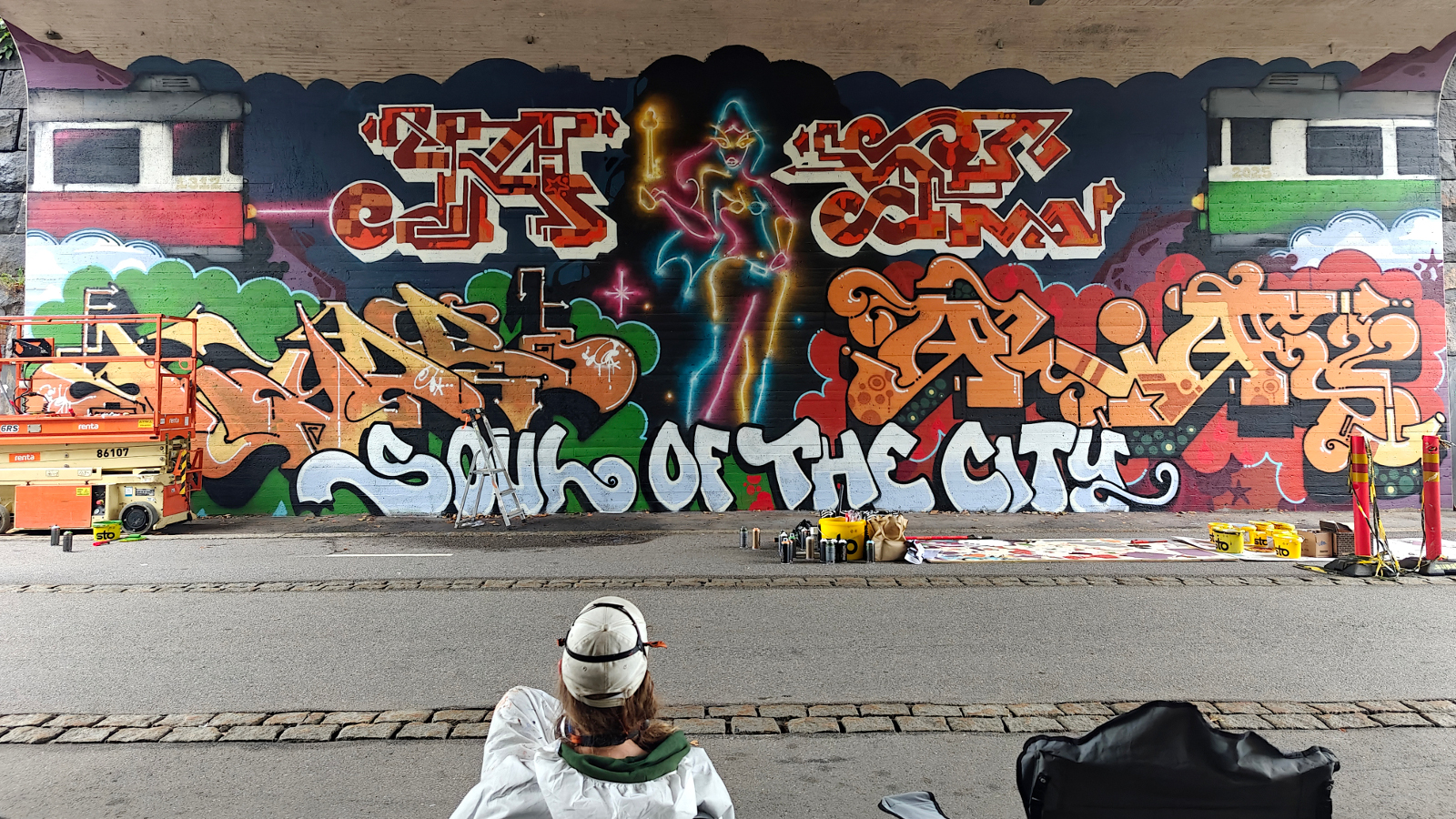 Kuvassa on graffitia taustalla olevalla betoniseinällä sillan alla.  Edessä on katselija valkoisessa kypärässä ja takissa. Välissä on harmaata asfalttia.
