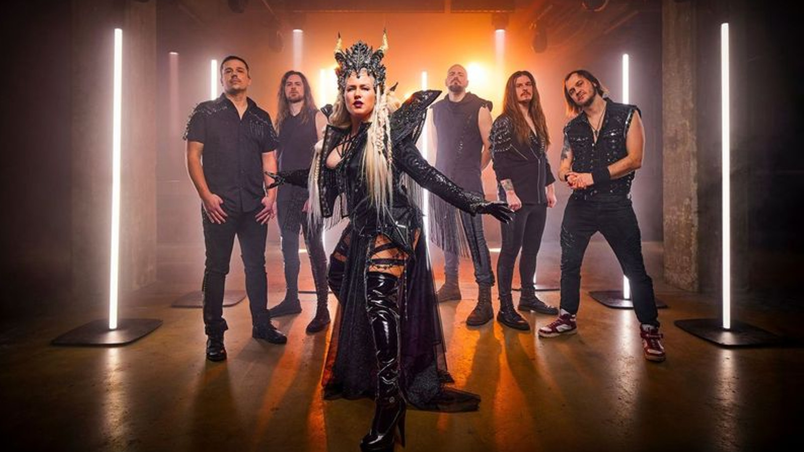 Kuvassa on Battle Beast ja etualalla on naissolisti mustassa näyttävässä puvussa, jossa on korkeat olkapääsiivet ja koristeellinen päähine sarvineen. Taustalla on viisi miest mustissa vaatteissa. Takana pystysuuntaisia led-valoja.