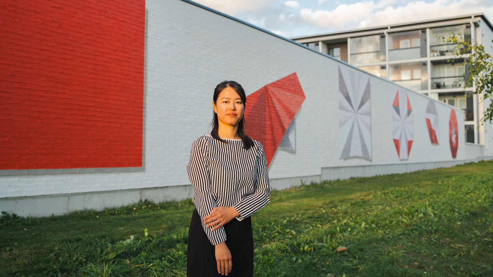 Kuvassa on pitkää seinämää ja siinä on vasemmala on maalattu punainen iso neilö. Vaalealla seinämällä on punainen origami sekä neljä muuta kuviota, joissa on myös harmaata. Edessä on seisomassa tekijä Momo Yoshino.