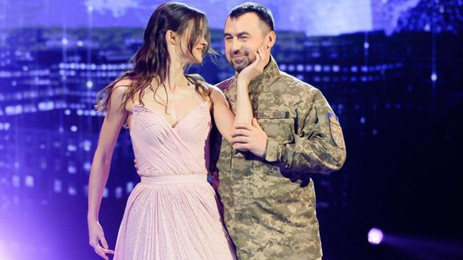 Kuvassa ovat Polina Ichenko roosassa avonaisessa tanssimekossa ja Artem Moroz armeijan takissa. Taustalla on violetti kaupunkimaisema kulissi.