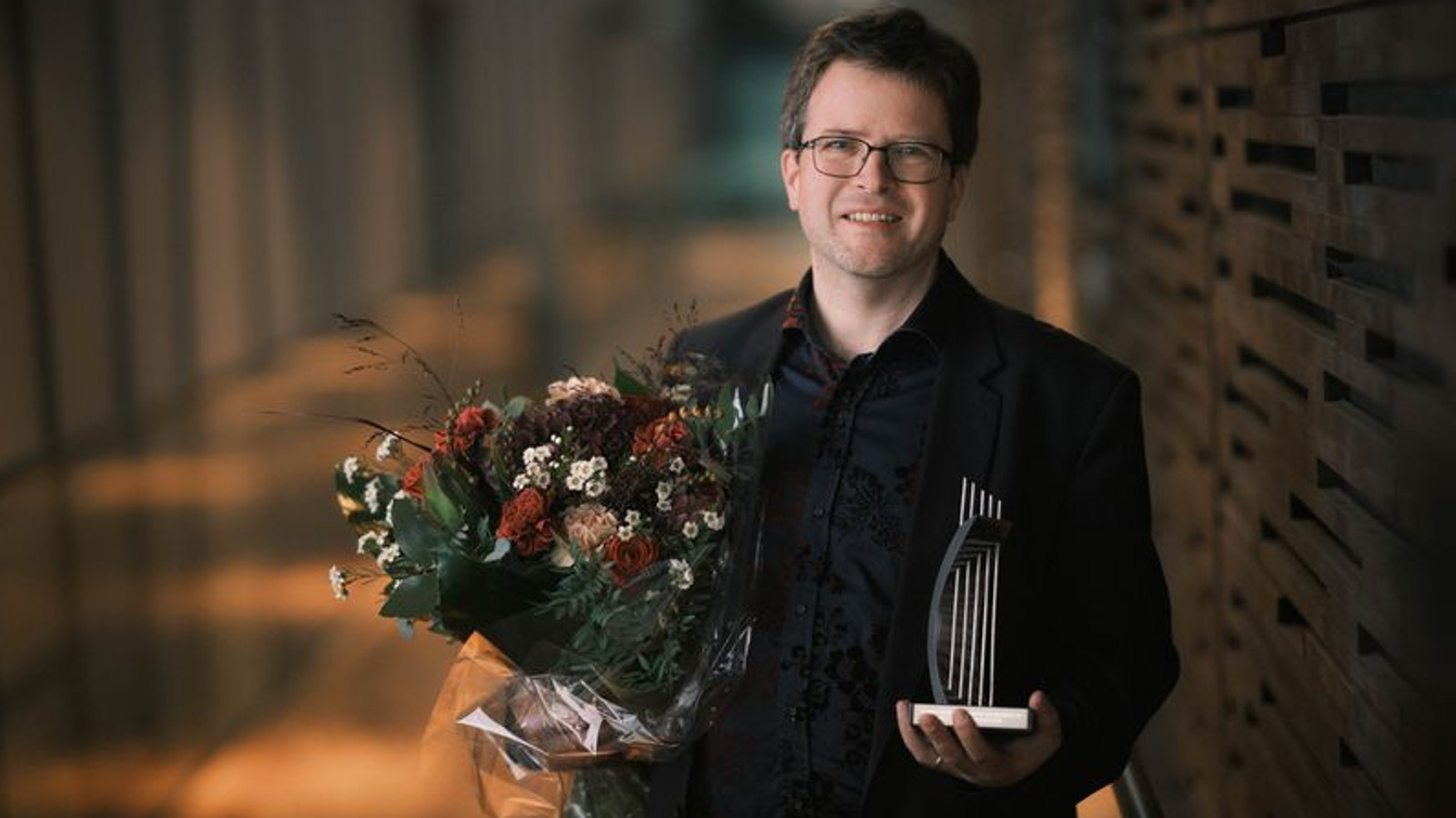 Kuvassa on Nils Scweckendiek puolivartalokuvassa ja hänellä on musta takki päällään. Vasemmassa kädessään hänellä on kukkakimppu ja oikeassa kädessään palkintoveistos, jossa on kaari ja nuottirivistö ylöspäin.