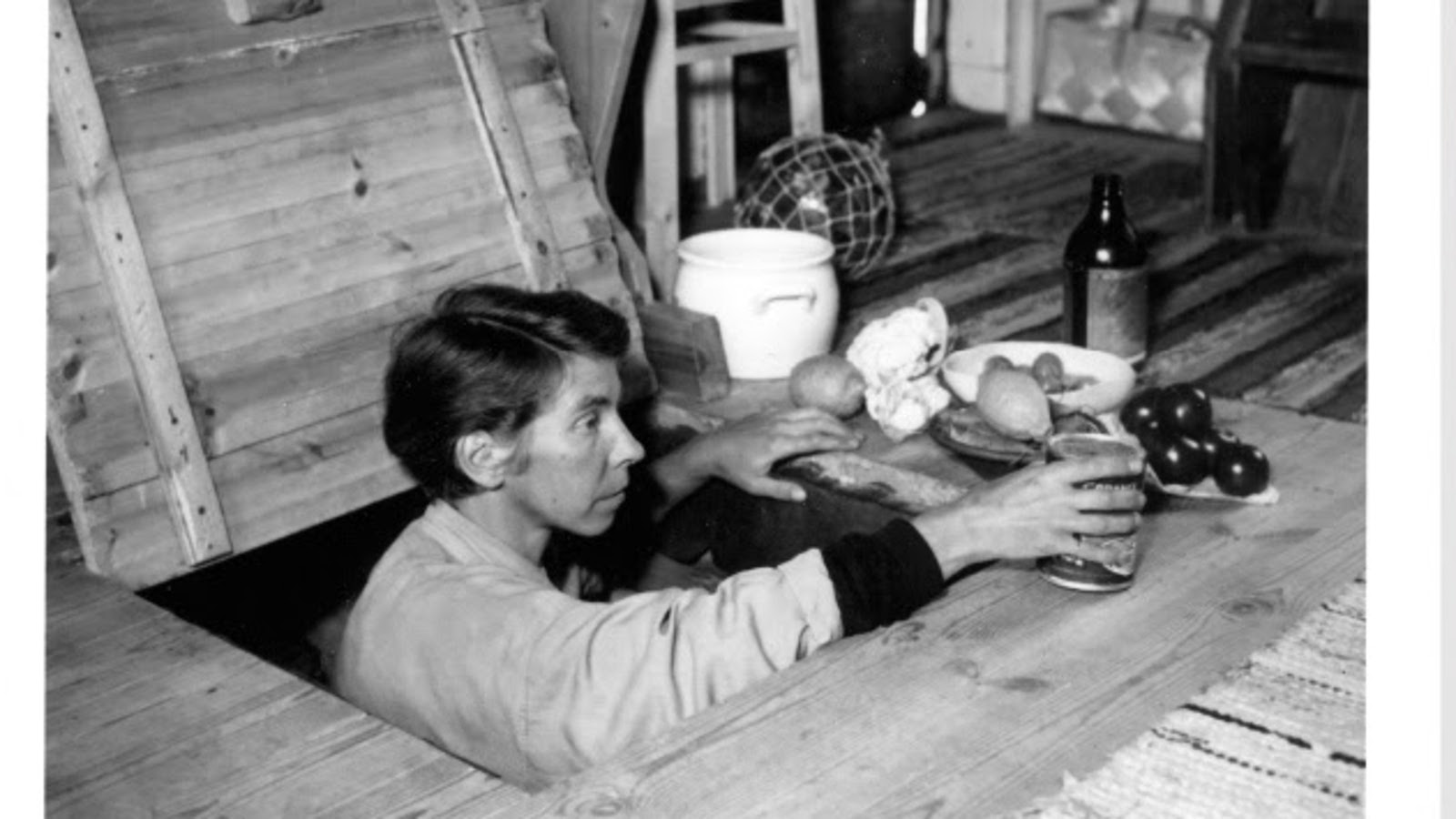 Kuvassa on Tove Jansson lattialuukussa ja hänen edessään lattialla on erilaista ruokaa lautasilla. Oikeassa kädessään hän pitää säilyketölkkiä. Kuva on musta-valkoinen.