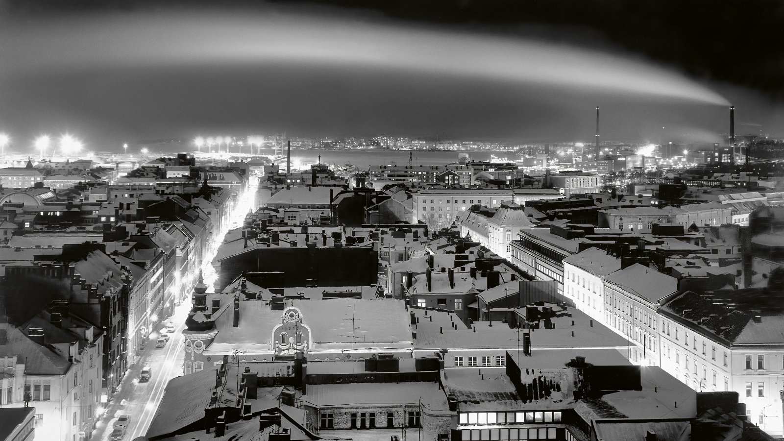 Kuvassa on yläviistosta näkymä talvisen, yöllisen Helsingin keskustan yli. Pimeällä taivaalla näkyy kaareva savuverho. Kuva on musta-valkoinen.
