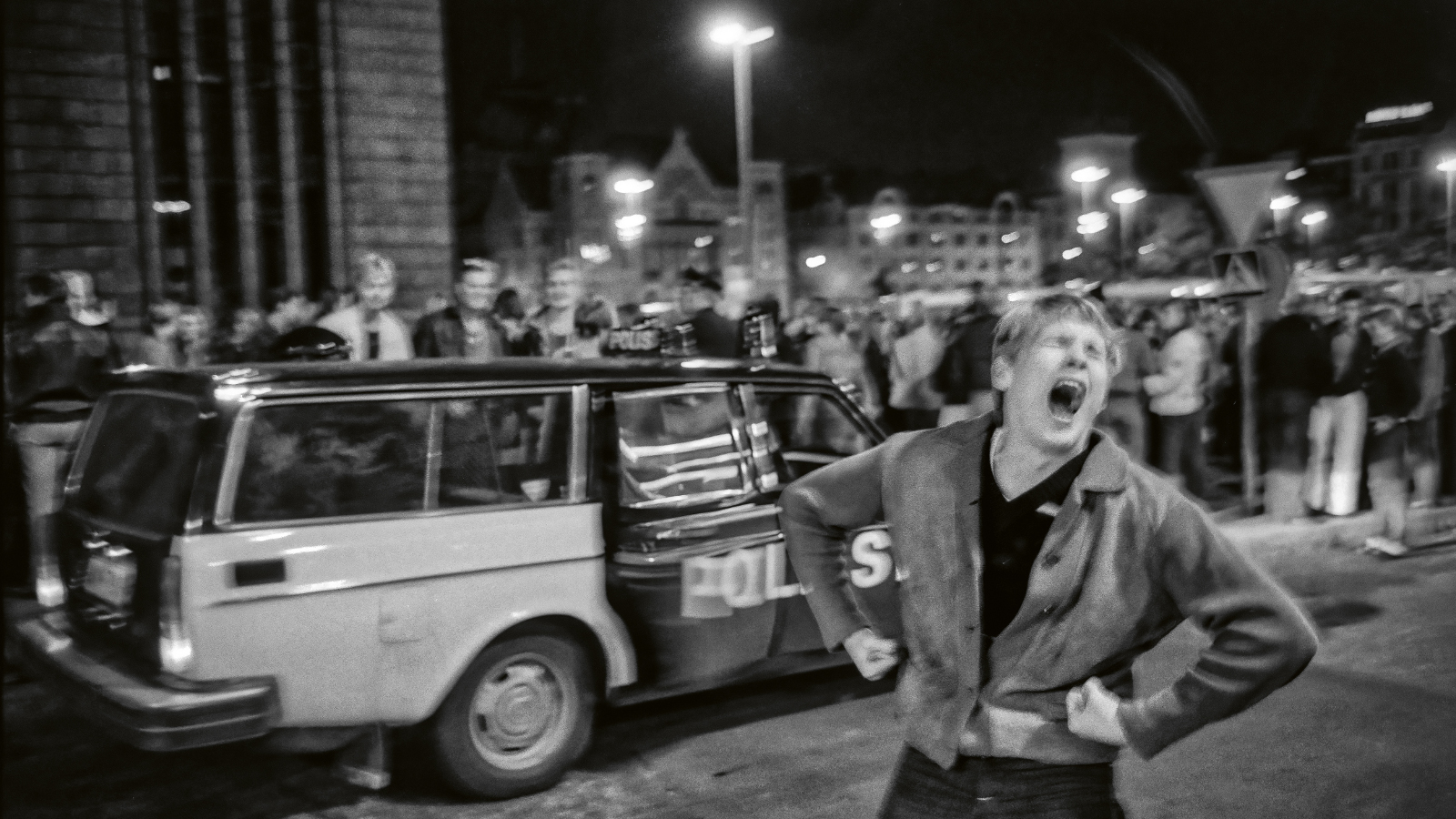 Kuvassa on edessä keskellä nuori mies pusakassa ja hän huutaa. Takana on poliisiauto ja sen takana nuorisojoukkoa Helsingin päärautatieaseman edessä yöllä. Kuva on musta-valkoinen.