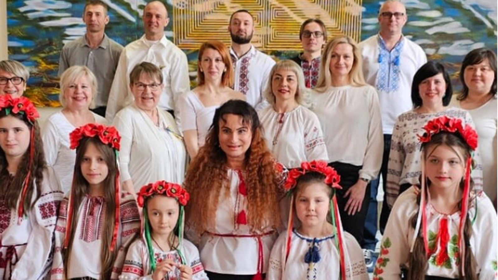 Kuvassa on kolmessa rivissä kuorolaisia ja edessä alhaalla on tyttöjä, seuraavassa rivissä naisia ja takarivissä miehiä. Heillä on valkoiset puserot tai kansallispuvun kirjaillut puserot. 