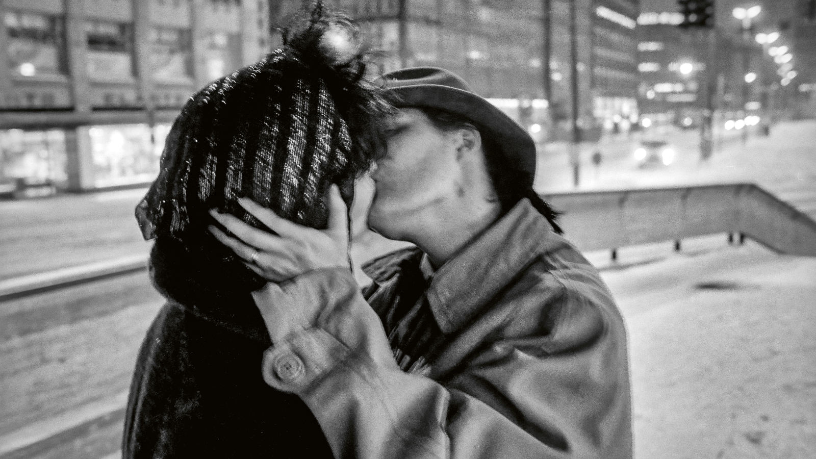 Kuvassa on puolivartalokuvassa nuori nainen ja mies suutelemassa. Nainen on puoliksi selin ja miehen kasvot näkyvät suudellessa. Miehellä on vaalea pusakka ja hän pitää vasenta kättään naisen niskassa. Taustalla näkyy talvista Hakaniemeä. Kuva on musta-valkoinen.