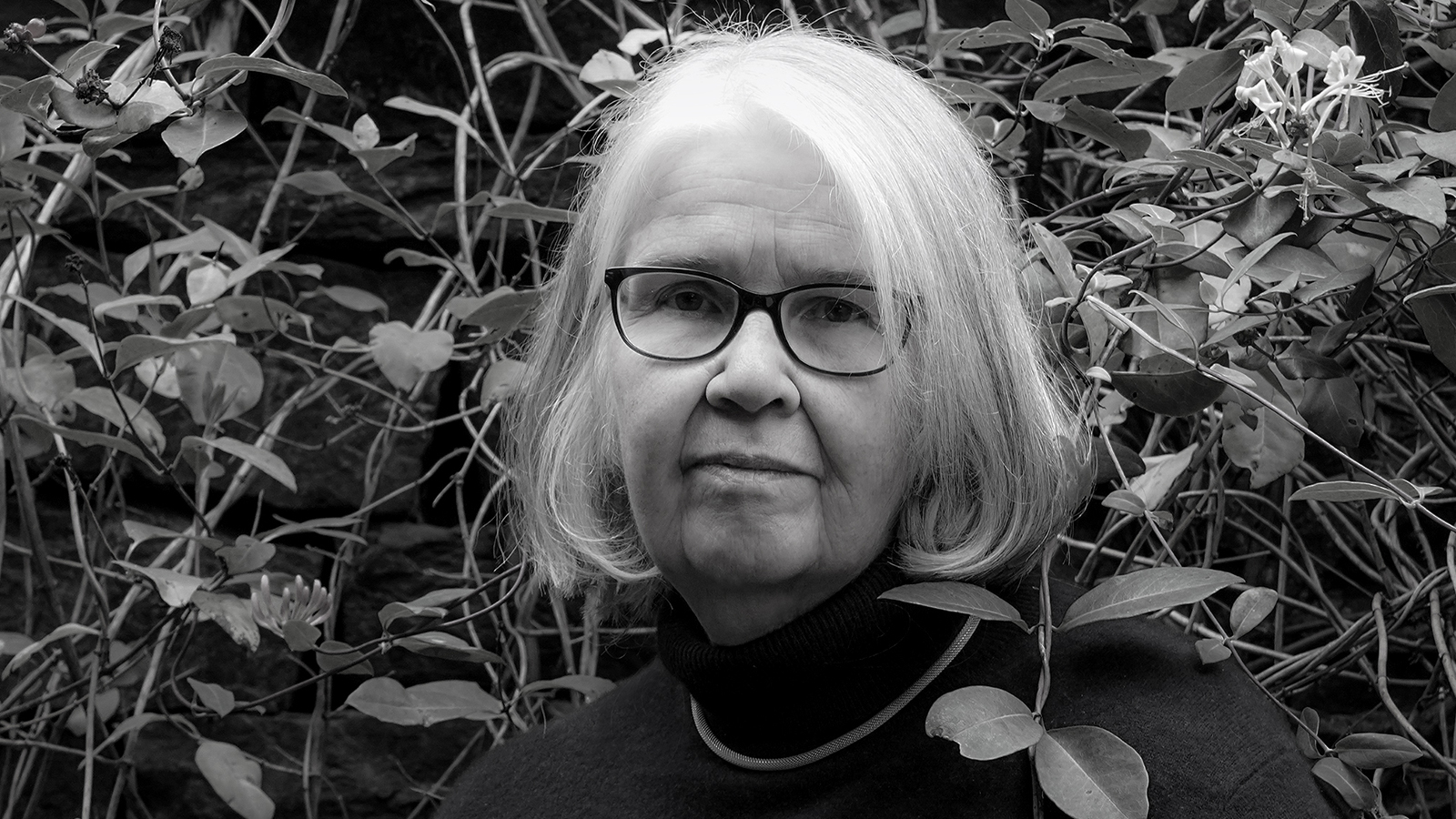 Kuvassa on Mona Schalin kasvokuvassa ja hänellä on vaaleat olkapäälle kaartuvat hiukset. Taustalla on lehteviä kasveja. Kuva on musta-valkoinen. 