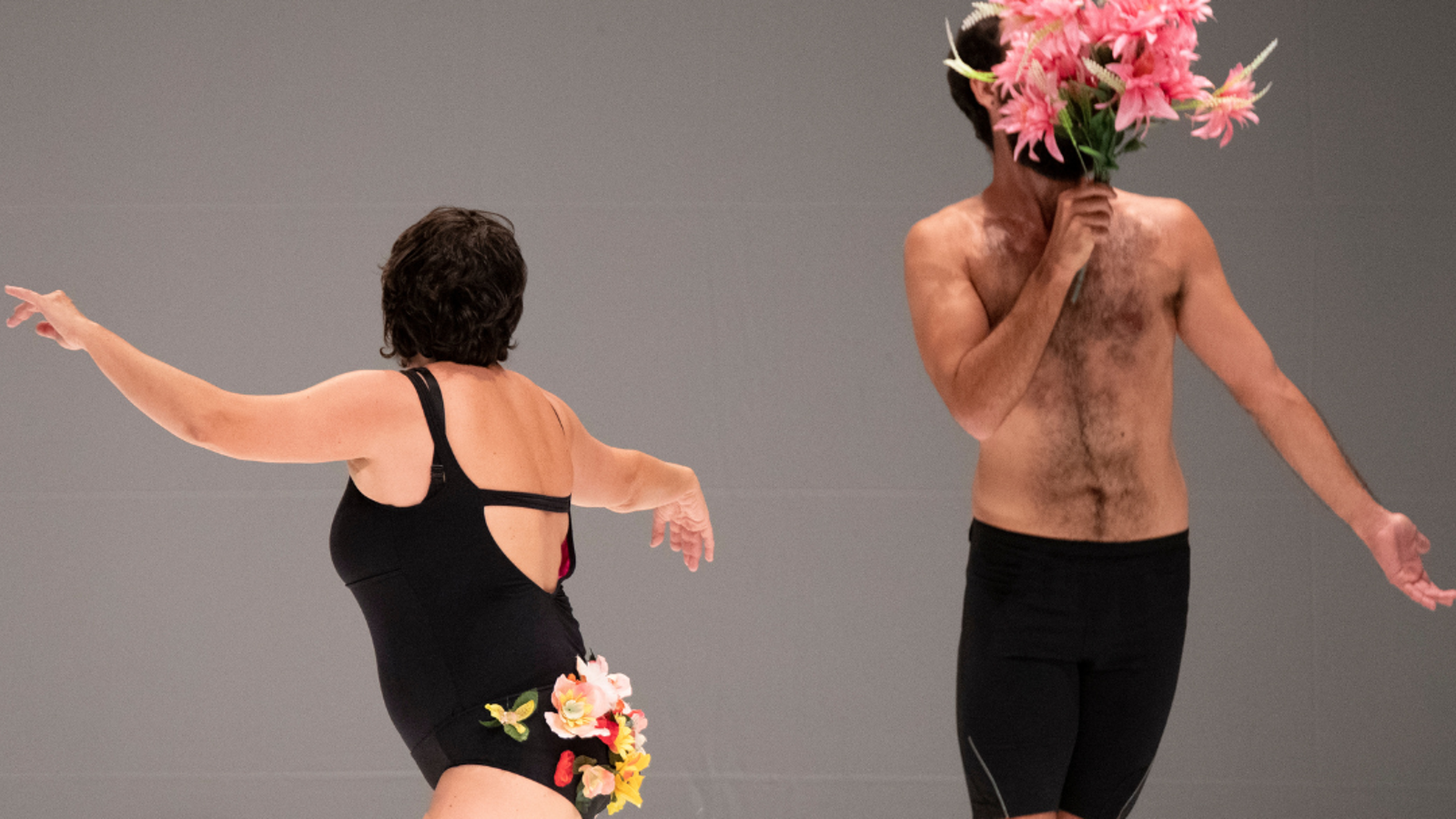 Kuvassa on kaksi miestanssijaa, joista vasemmalla olevalla on musta uimapuku päällään ja siinä takapuolen kohdalla kukkia. Oikean puoleisella on paljas yläruumis ja kukkia kasvojen edessä.