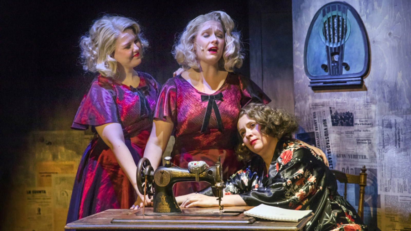 Kuvassa on edessä vanhanaikainen poljettava ompelukone ja sen takana on oikealla tummahkossa mekossa nainen istumassa. Basemmalla ompelukoneen ääressä kaksi violetin punaisissa mekoissa kaksi vaalean kiharatukkaista naista laulamassa.