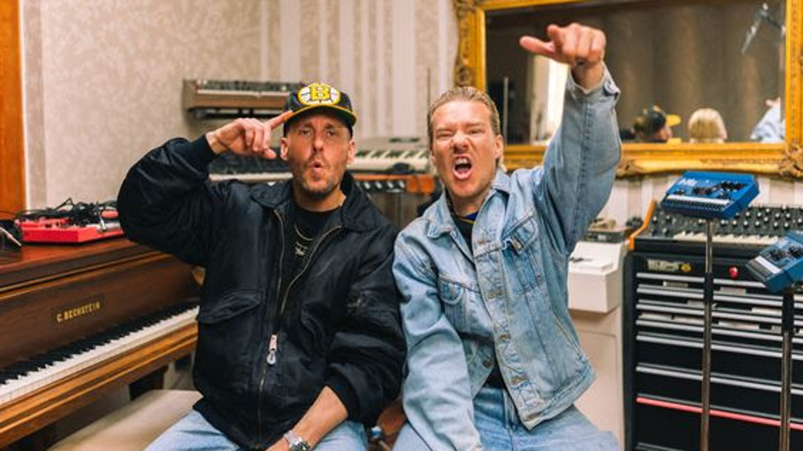 Kuvassa ovat JVG jäsenet ja vasemmalla Ville Gallella on musta nahkatakki ja Jarella vaalean sininen farkkupuku sekä vasen käsi ylhäällä. Taustalla on on soittimia ja äänitysstudio.