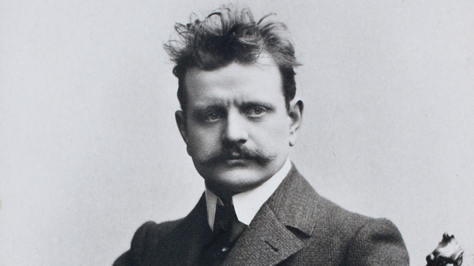Kuvassa on  Jean Sibelius puolivartalokuvassa ja hänellä on hiukset päälaella pörhöllään ja paksu takki päällään sekä valkoiset kaulukset. Kuva on musta-valkoinen.