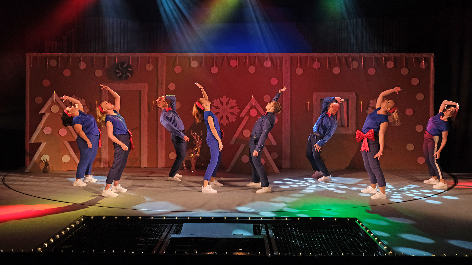 Kuvassa on kahdeksan henkilöä sinisissä asuissa taipuneina taaksepäin toinen käsi ojennettuna ylös. Taustalla on ruskeaa seinämää, jossa on ruskehtavia joulukuusia.