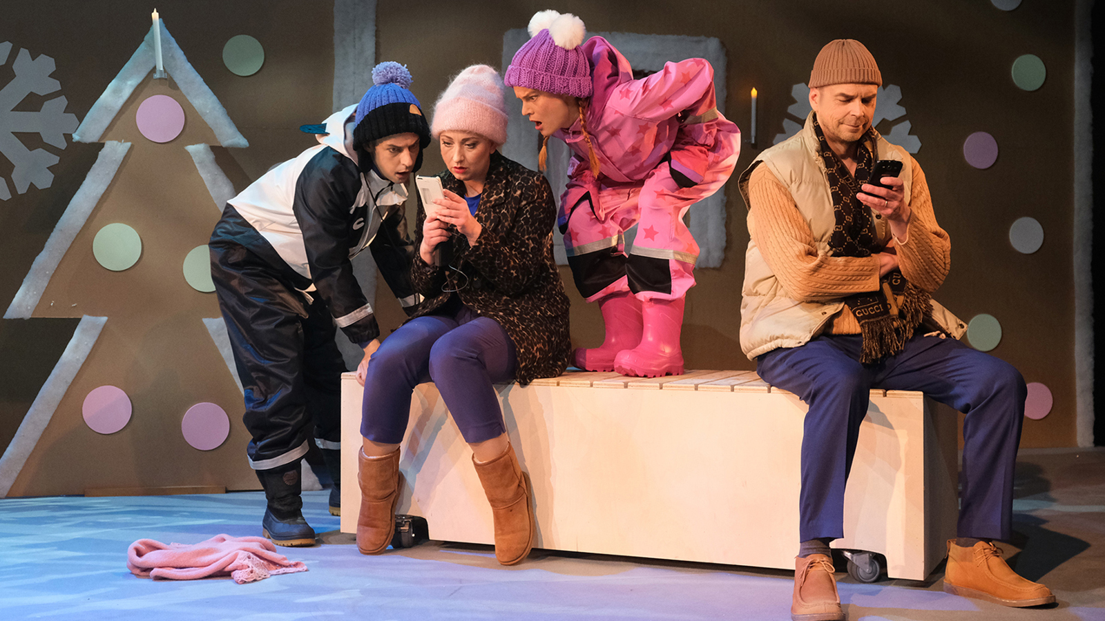 Kuvassa on ison penkin päällä istumassa vasemmalla farkuissa ja pusakassa katsomassa lapsia esittävien vasemmalla sini-valkoisissa vaatteissa sekä kumisaappaissa poika ja penkin päällä tyttö pinkeissä ulkovaatteissa katsomassa kännykkää yhdessä. Penkin päässä on ruskeassa pusakassa ja farkuissa mies katsomassa kännykkää