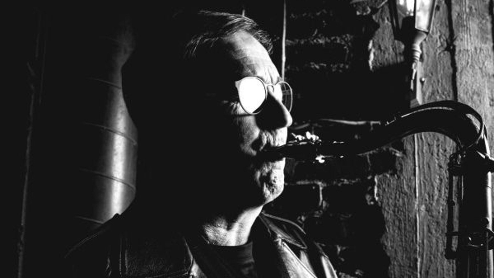 Kuvassa on Jussi Saksa soittamassa saksofonia puolivartalokuvassa. Kuva on tumma ja silmälasit kiiltävät ja kasvot näkyvät osittain.