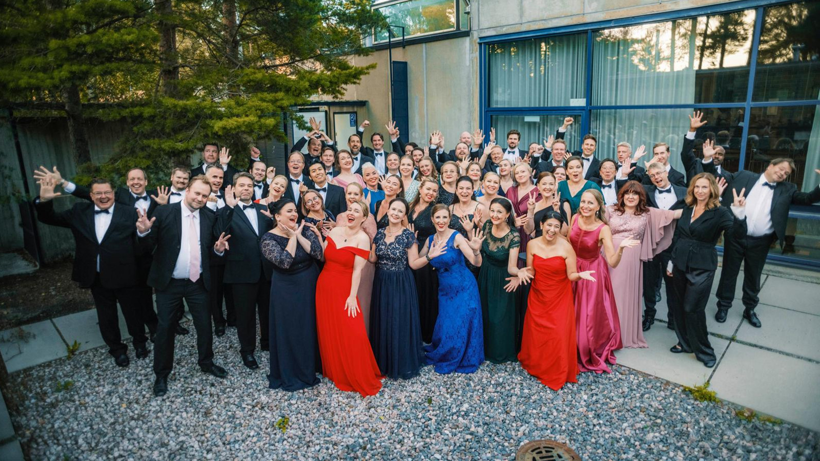 Kuvassa on koko kansallisoopperan kuoro seisomassa pihalla. Vasemmalla ja takana ovat miehet musta-valkoisissa puvuissa. Edessä ovat naiset pitkissä, eri värisissä pitkissä iltapuvuissa. 