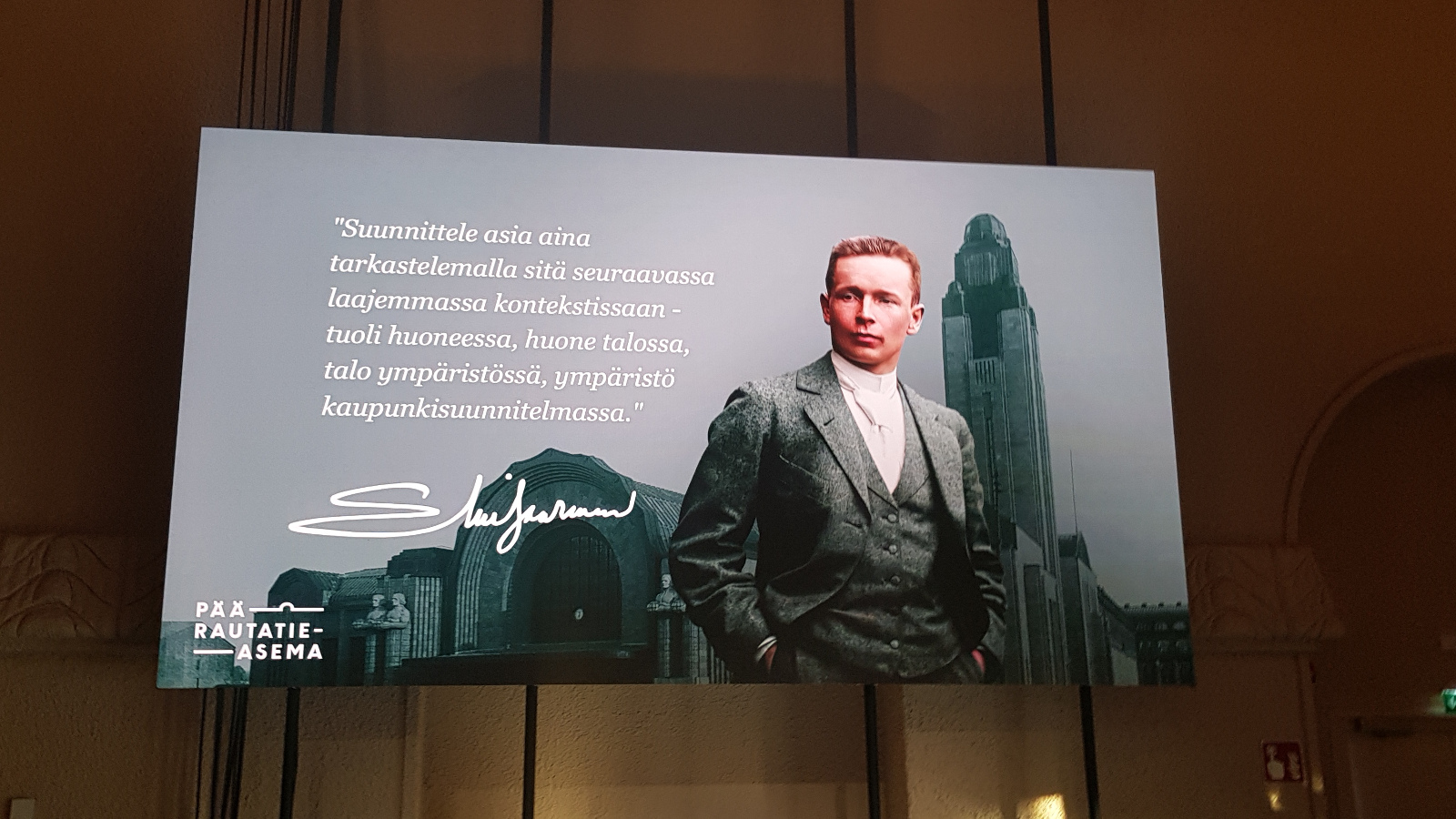 Kuvassa on taulu, jossa on tekstiä valkoisella ja vieressä seisoo nuori Eliel Saarinen harmaassa puvussa. Taustalla on Helsingin päärautatieasema taulun takana tummaa seinää.