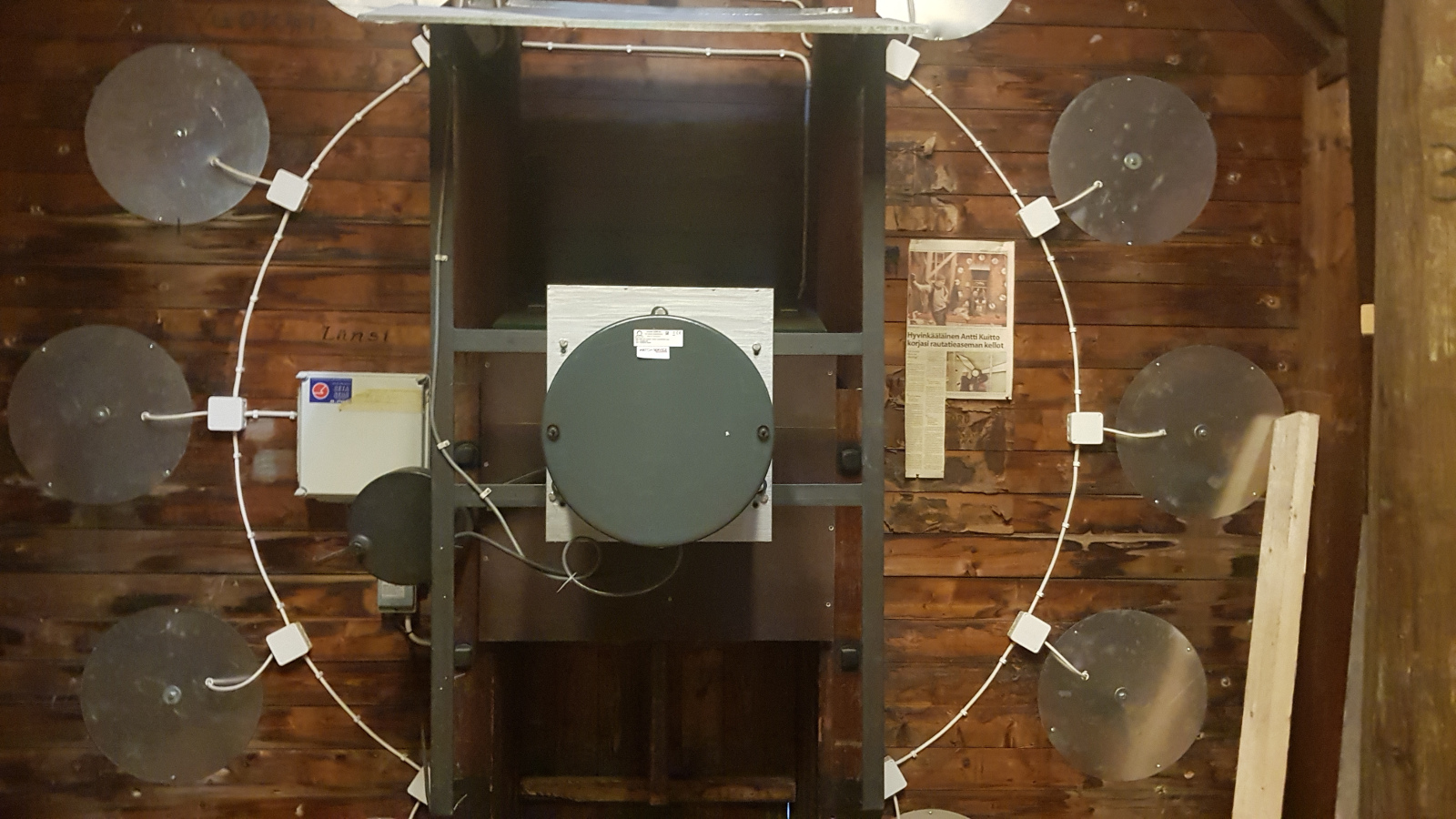 Kuvassa on aseman kellon pyöreä metallinen ohut iso rengas ja sen ympärillä on harmaat pyöreät lätkät. Tausta on ruskea.