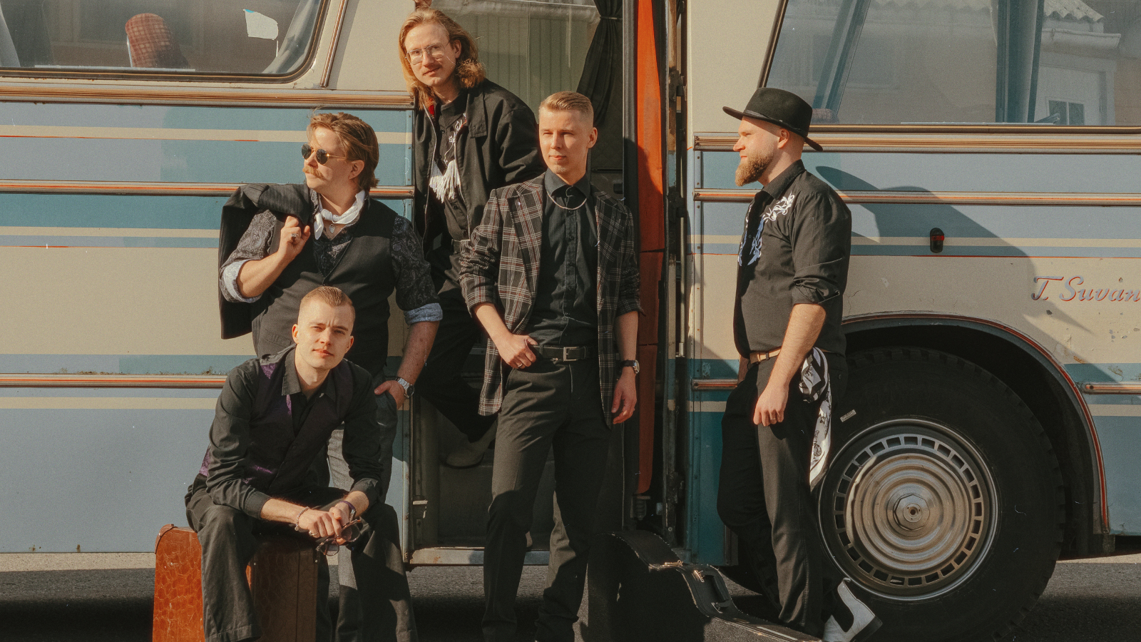 Kuvassa ovat Komiat-yhtyen viisi jäsentä ja he ovat vaalean sini-keltaisen bussin edessä. Neljä heistä seisoo ja yksi istuu matkalaukun päällä. 