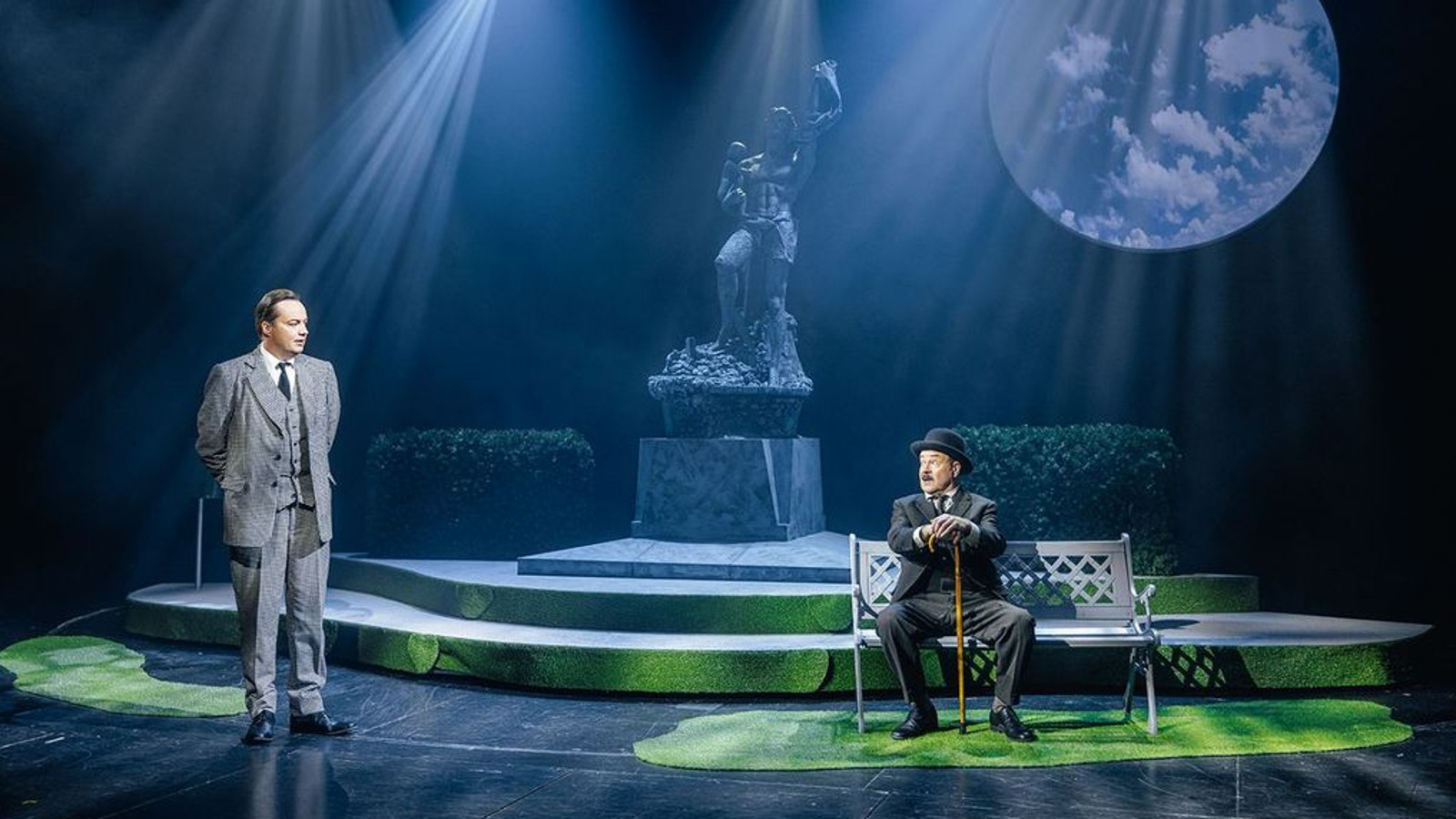 Kuvassa on vasemmalla seisomassa harmaassa puvussa mies ja oikealla puiston penkillä istumassa ruskeassa puvussa ja knallissa keppi kädessään, johon nojaa. Taustalla on Havis Amandaa muistuttava patsas. Taustalla on tumma iltavalaistus. Oikealla ylhäällä on suuri kuu.