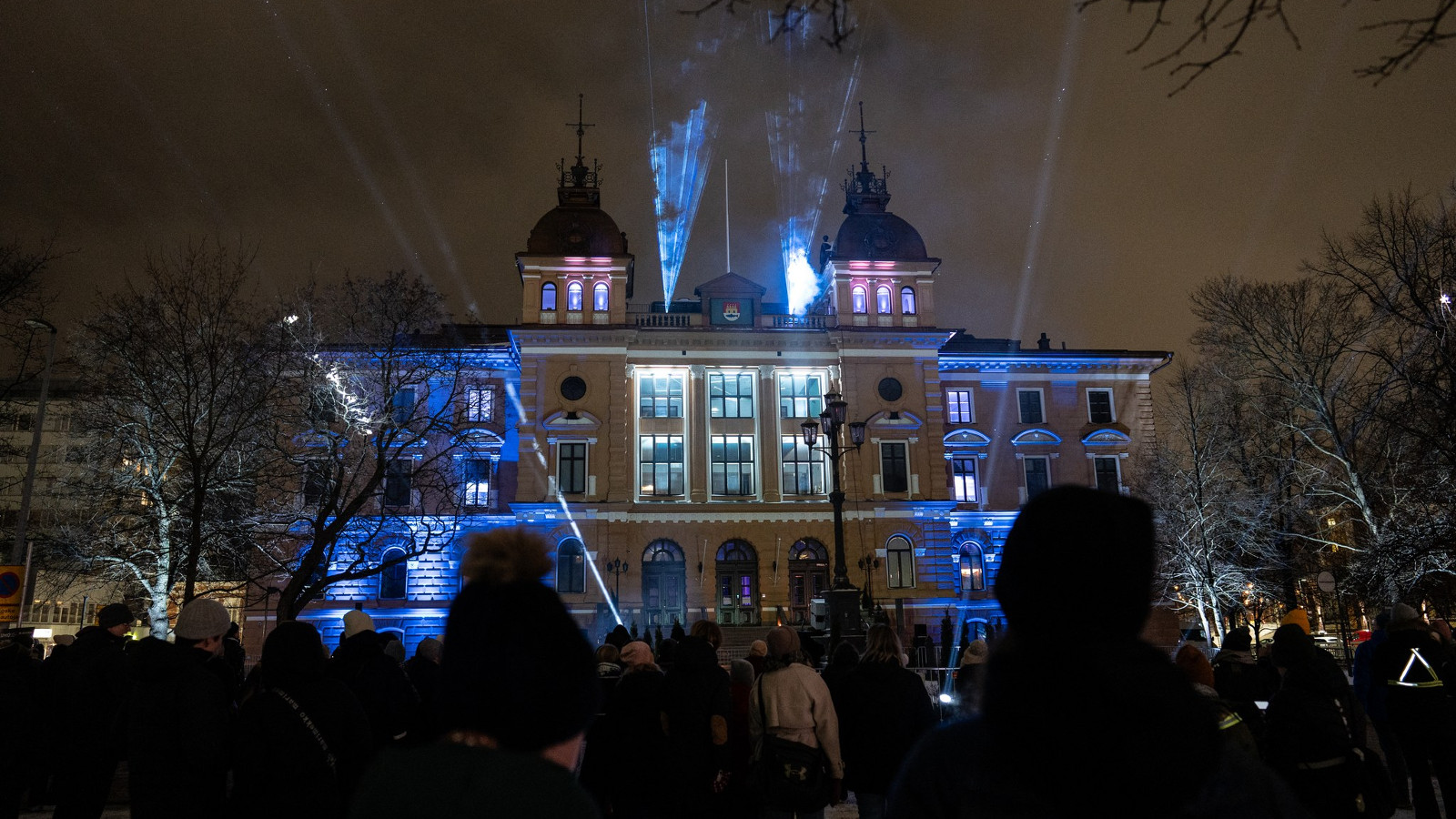 Kuvassa on Oulun kaupungintalo yöllä ja sitä valaistaan sinisillä valoilla molemmilta reunoilta. Talossa on kaksi tornia ja kolme korkeaa valaistua ikkunaa.