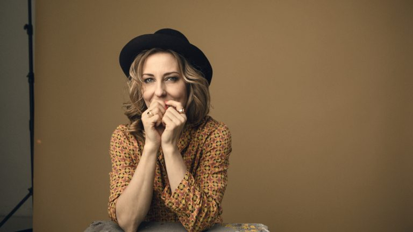 Kuvassa on Mariska puolivartalokuvassa ja hänellä on musta hattu päässään sekä kellertävä, pikkukuvioinen pusero päällään. Hänellä on molemmat kädet kevyesti nyrkissä suun edessä. Tausta on ruskehtavaa seinää.