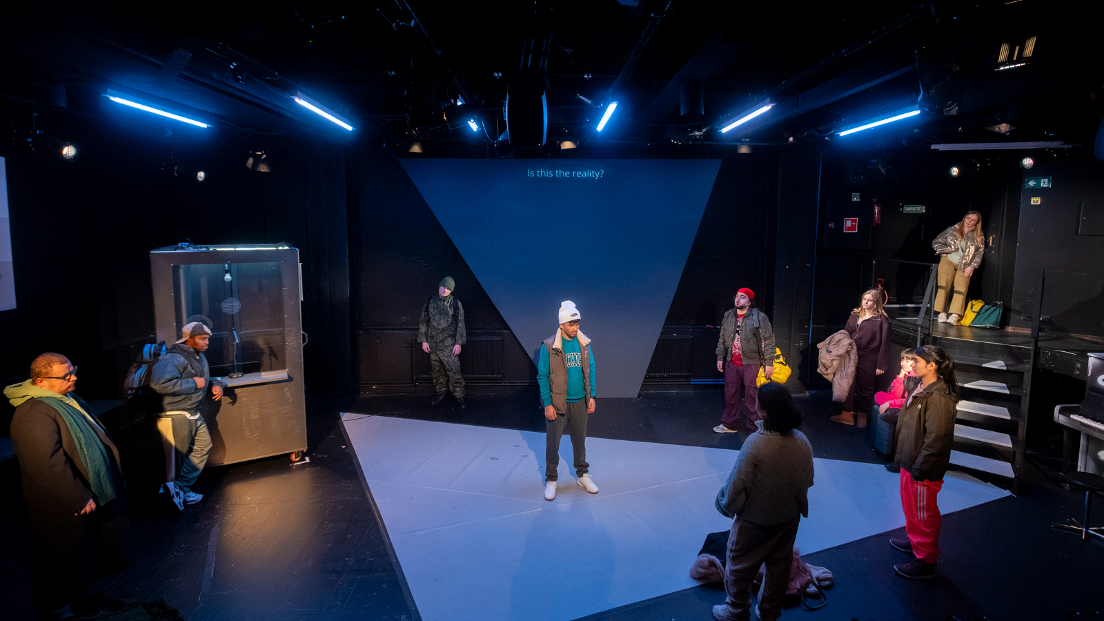Kuvassa on näyttämö, johon kohdistuu sinistä valoa. Keskellä seisoo nuori mies talvivaatteissa.  Ympärillä kauempana on joka puolella näyttämön reunalla nuoria ulkovaatteissa.