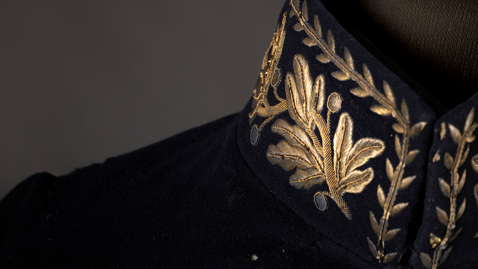 Kuvassa on mustassa takissa hieman olkapäätä ja pystykauluksen puolikas, jossa on kultakirjailua. Tausta on tumma.