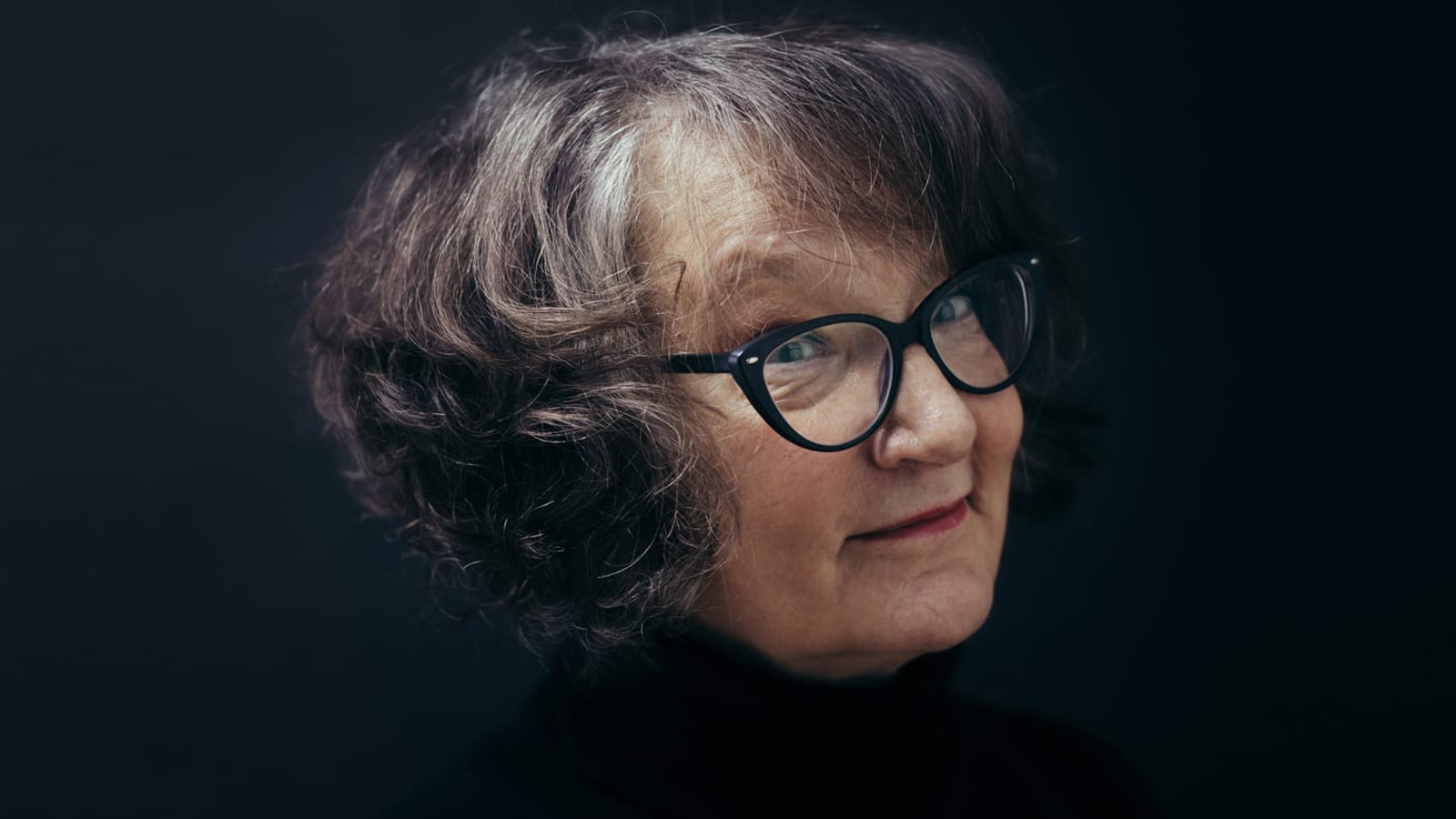 Kuvassa on Monika Fagerholmin kasvokuva ja hänellä on kihartavat tumman harmaat hiukset  sekä mustasankaiset silmälasit. Tausta on musta.