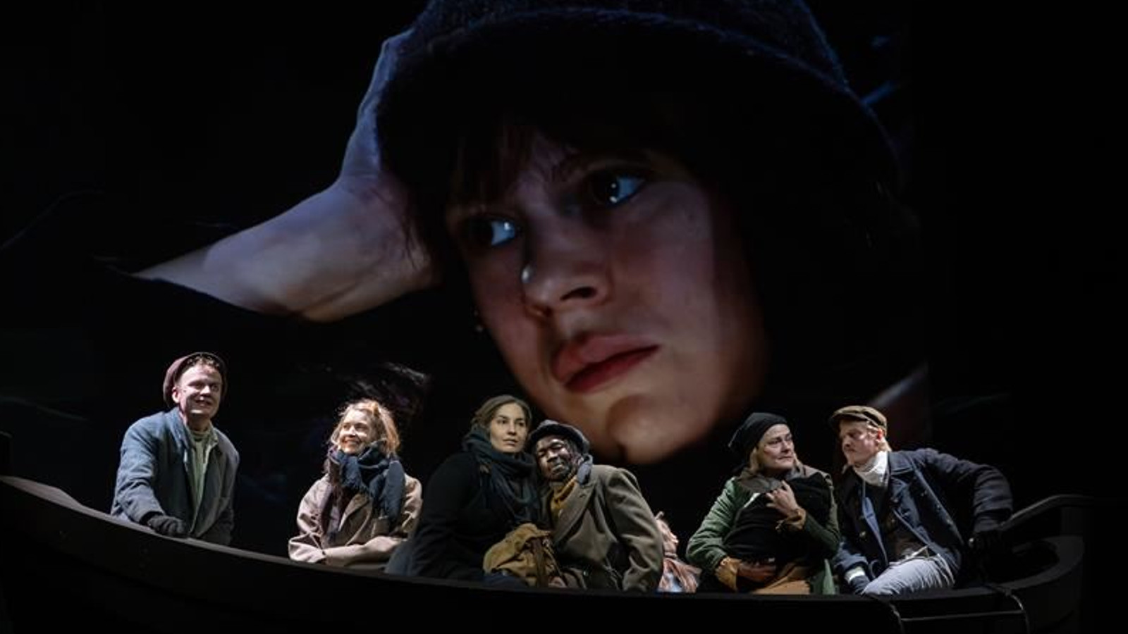 Kuvassa on veneessä kuusi henkilöä tummahkoissa vaatteissa. Taustalla on tummalla taustalla nuoren naisen kasvokuva lierihatussa.