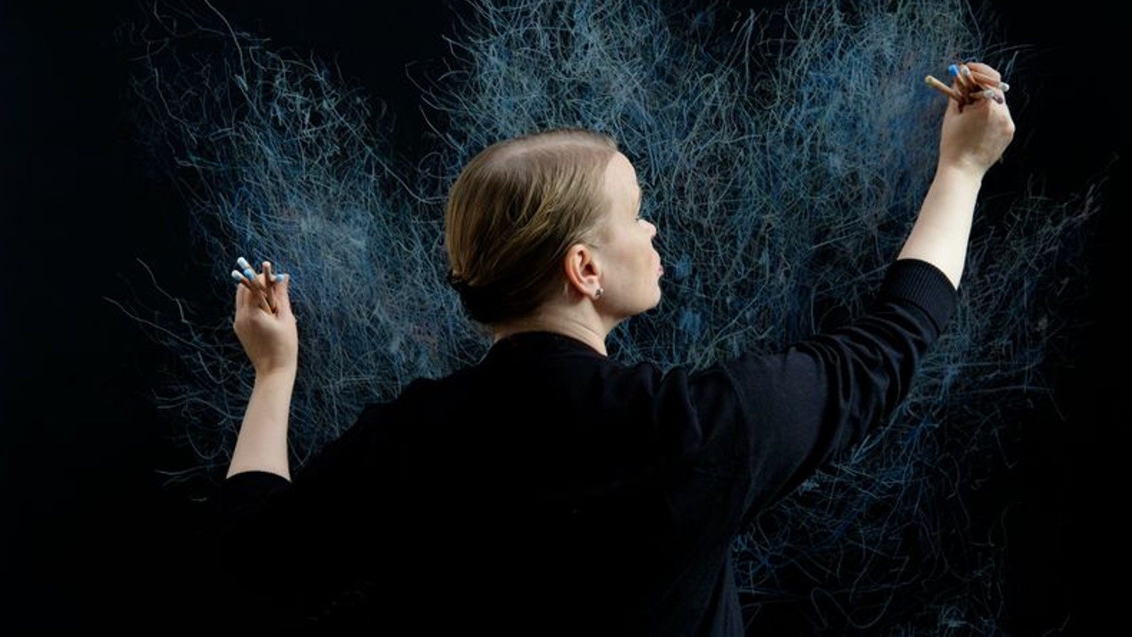 Kuvassa on Varpu Rossi piirtämässä tummalle seinälle puolivartalokuvassa kädet levitettyinä maalausasentoon. Hänellä on musta pusero.